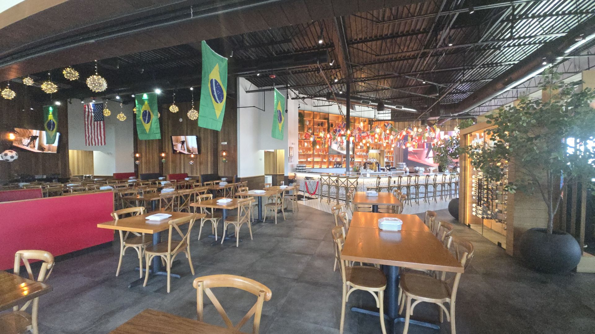 Spacious restaurant interior with wooden tables and chairs, Brazilian flags hanging from the ceiling, warm pendant lights, a long bar with stools, TV screens, and lush plants along the right.