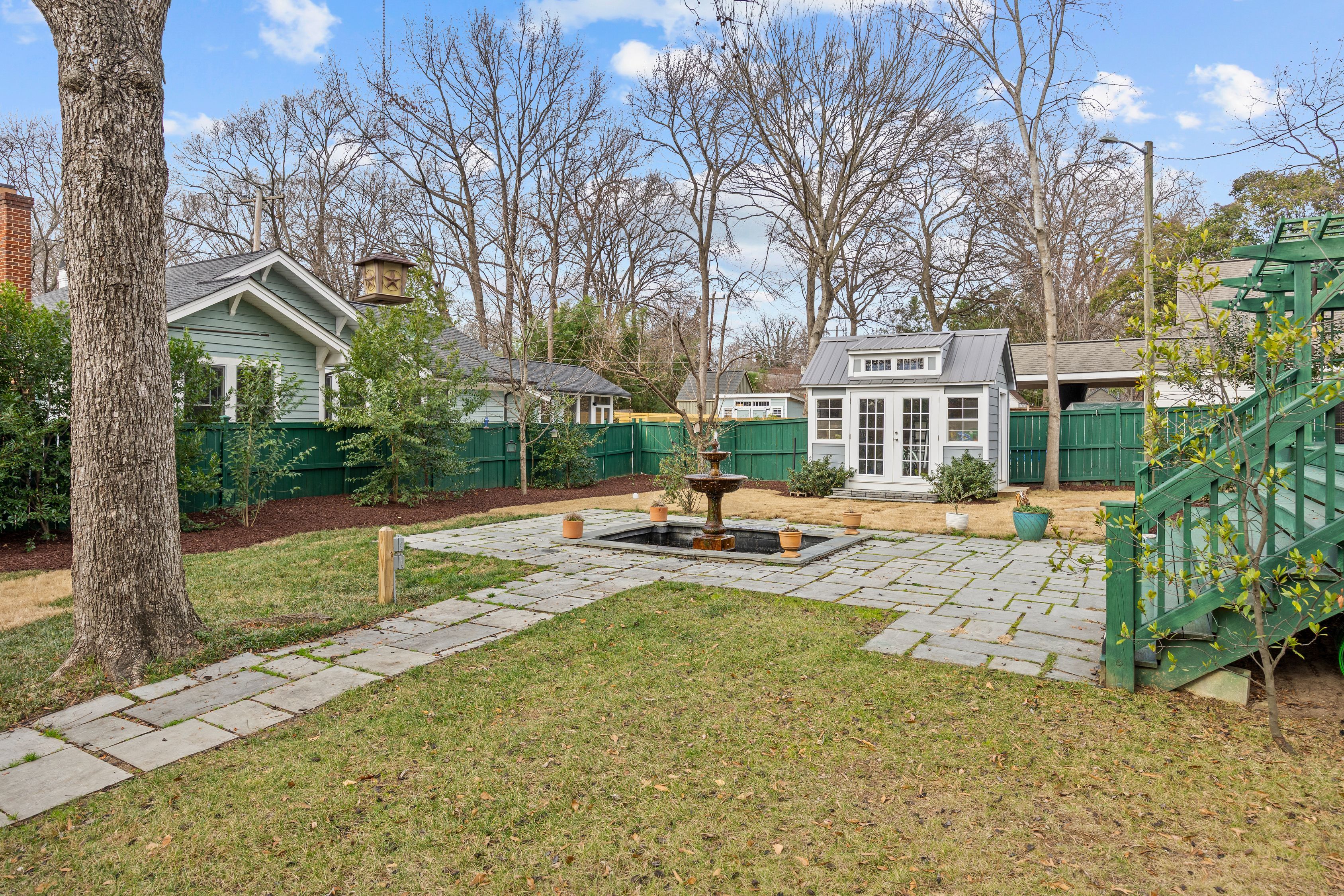 Backyard with green grass, stone paths, a brown fountain in the center, potted plants, green wooden fence, leafless trees, and a small white shed with glass doors under a blue sky.