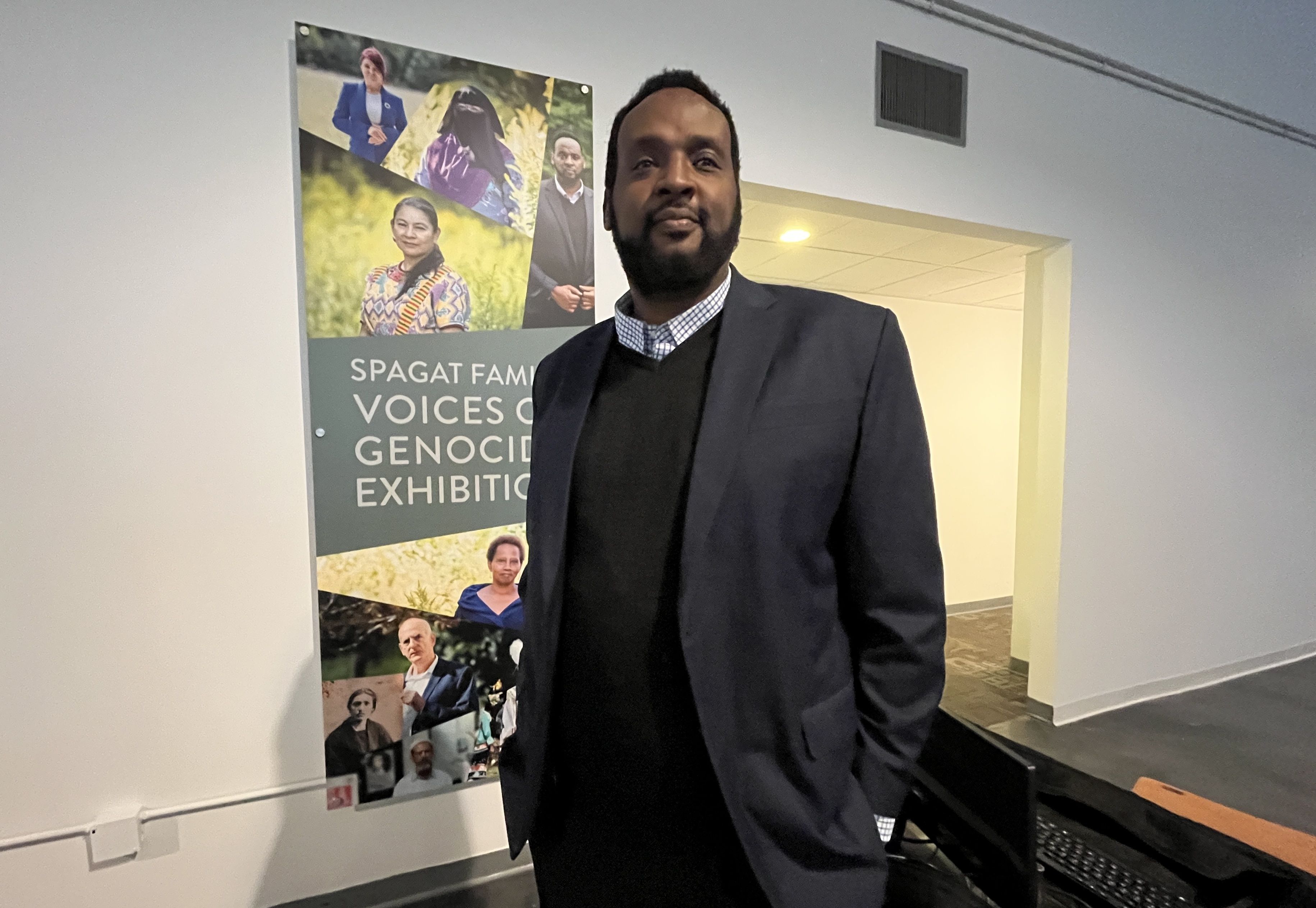 Kizito Kalima in a dark blazer and sweater stands indoors near a wall with a banner reading "Spagat Family Voices of Genocide Exhibition" featuring portraits of diverse people.