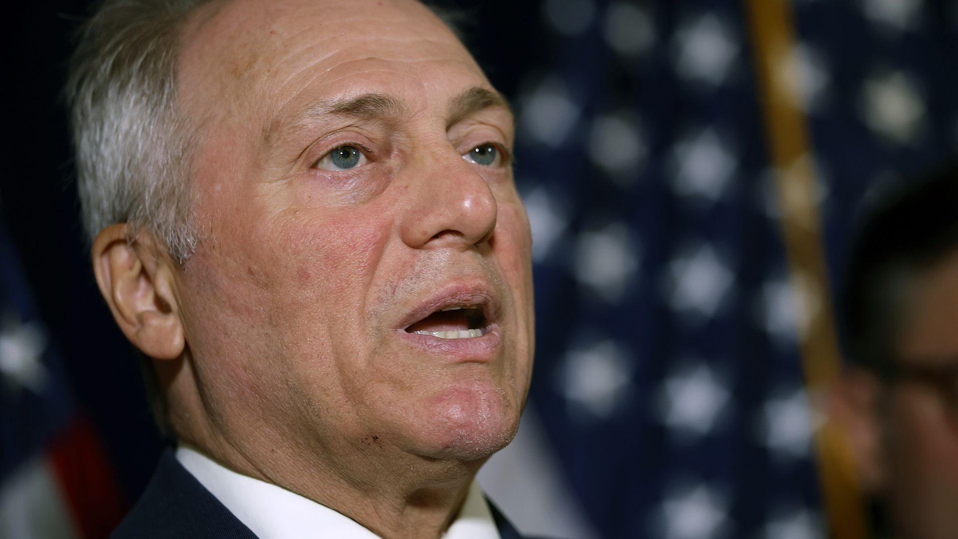 Close-up of a middle-aged man speaking, wearing a dark suit and red tie, with a blurred American flag background.