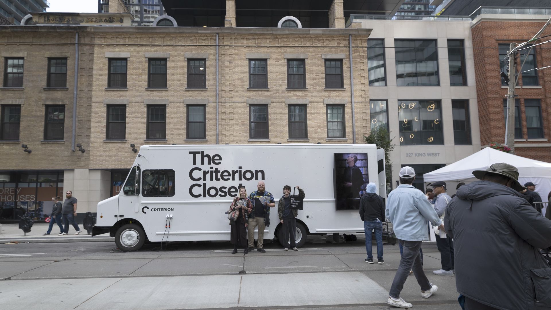 City street scene with a white trailer reading "The Criterion Close" parked before brick buildings; a small crowd stands nearby as modern glass buildings and a white tent rise in the background.