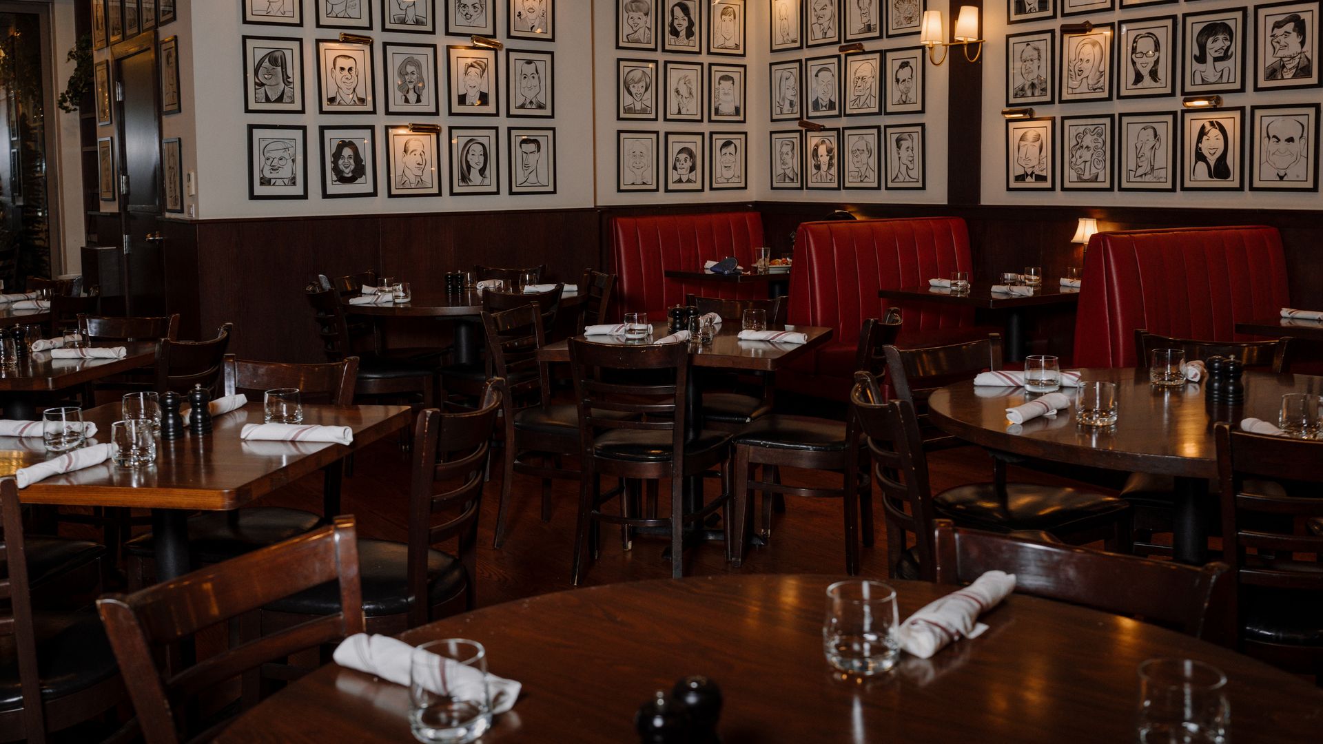 A restaurant dining room with cream walls lined by numerous framed black-and-white caricature portraits, red booth seating, dark wooden tables, and warm wall lighting.