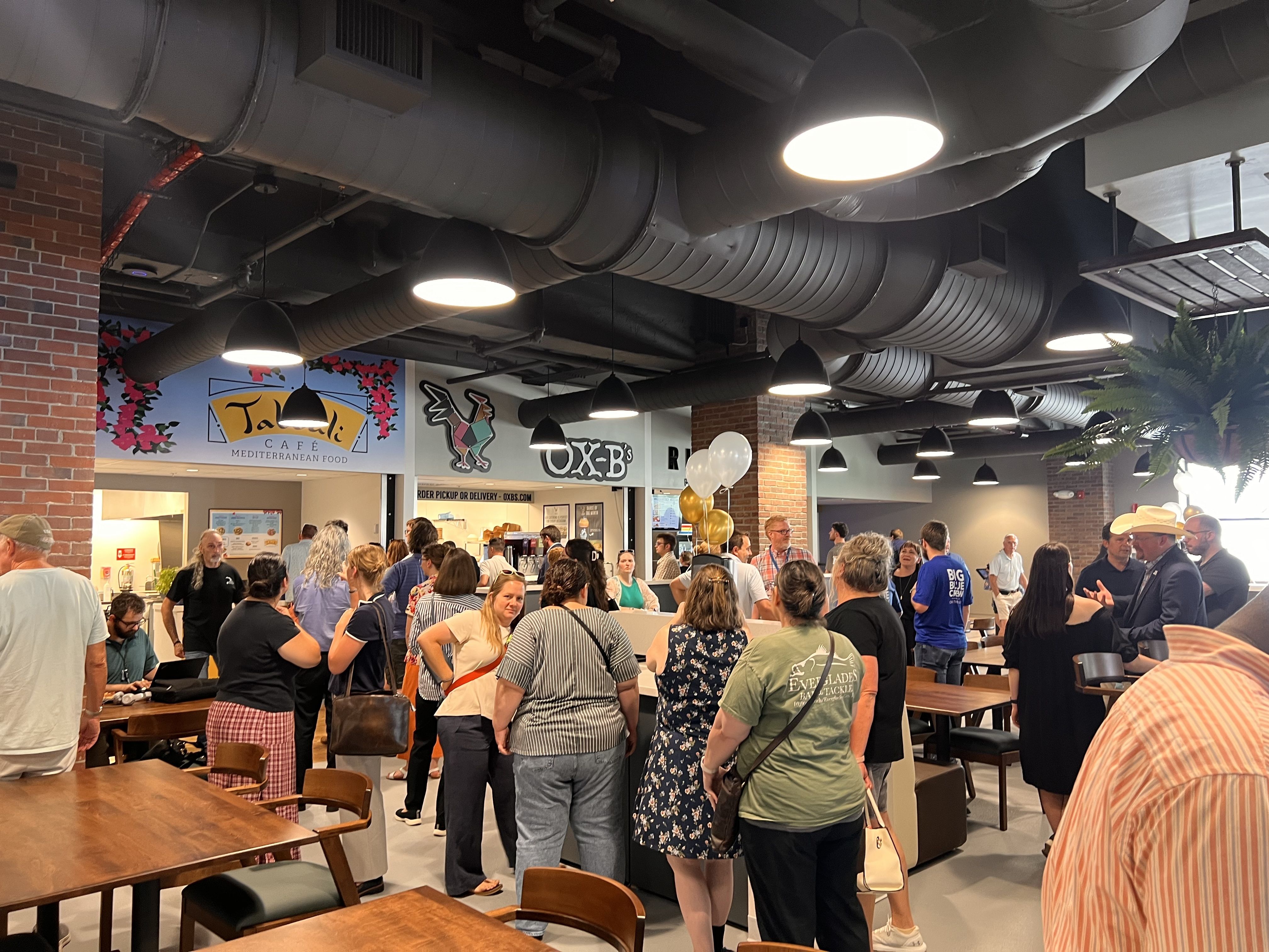Busy indoor food hall with large exposed gray ducts and round hanging lights. Crowds gather near a cafe counter beneath brick pillars, posters, and balloons, with wood tables and chairs.