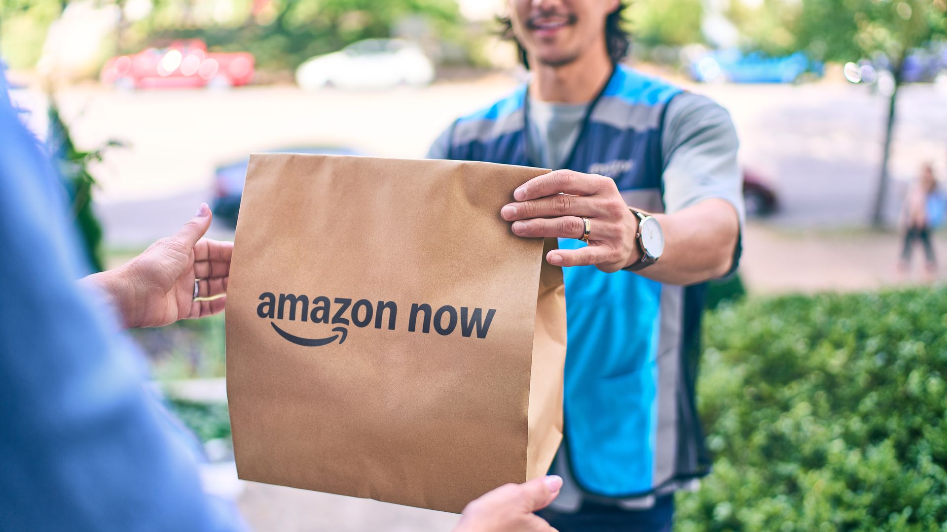 Delivery worker in a blue vest hands a brown paper bag labeled amazon now to a customer outside, with green trees and a street blurred in the background.