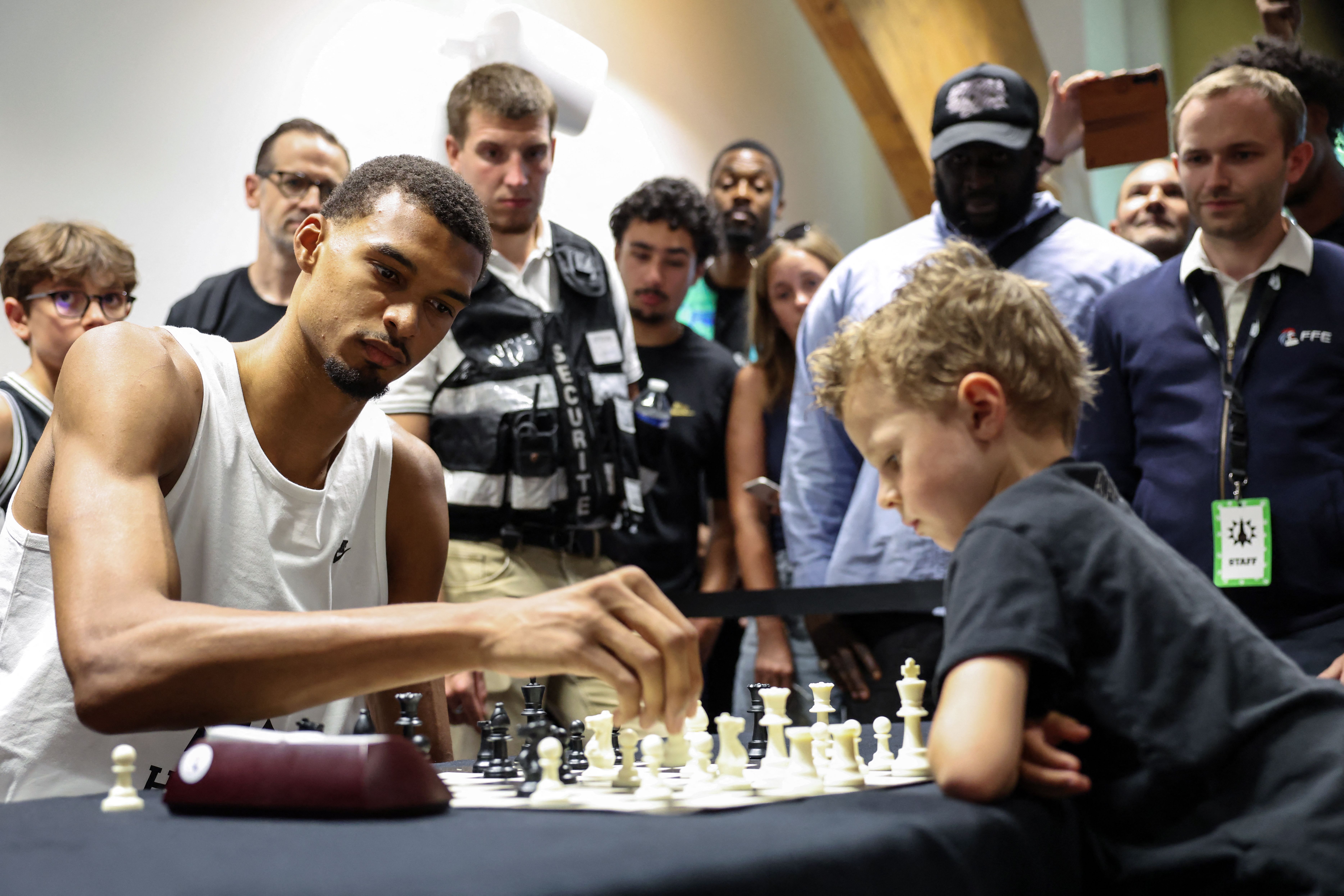 A young man in a white sleeveless shirt plays chess at a table, focused on the board. A crowd of onlookers, some with staff badges, watches intently in a busy room.