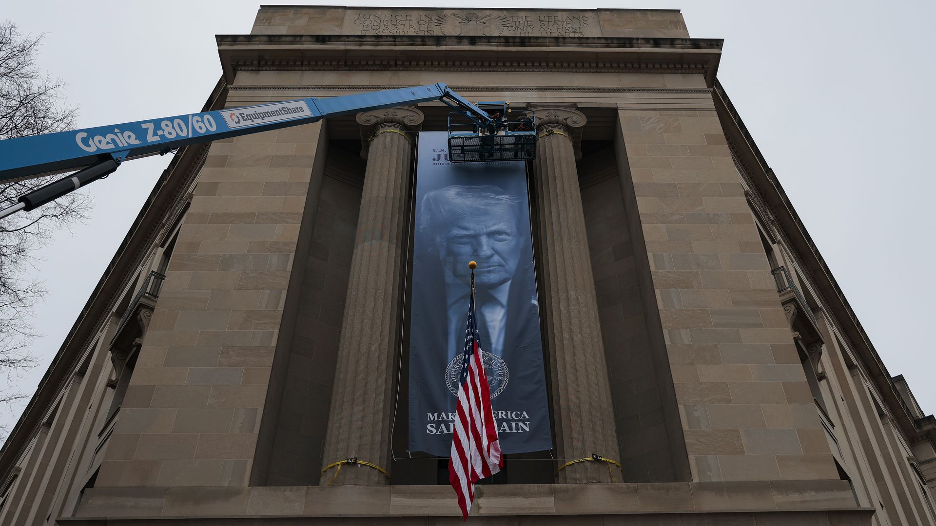 A large blue banner with a portrait of Donald Trump hangs between stone columns on a building, with an American flag in front and a blue lift platform positioned at the top.