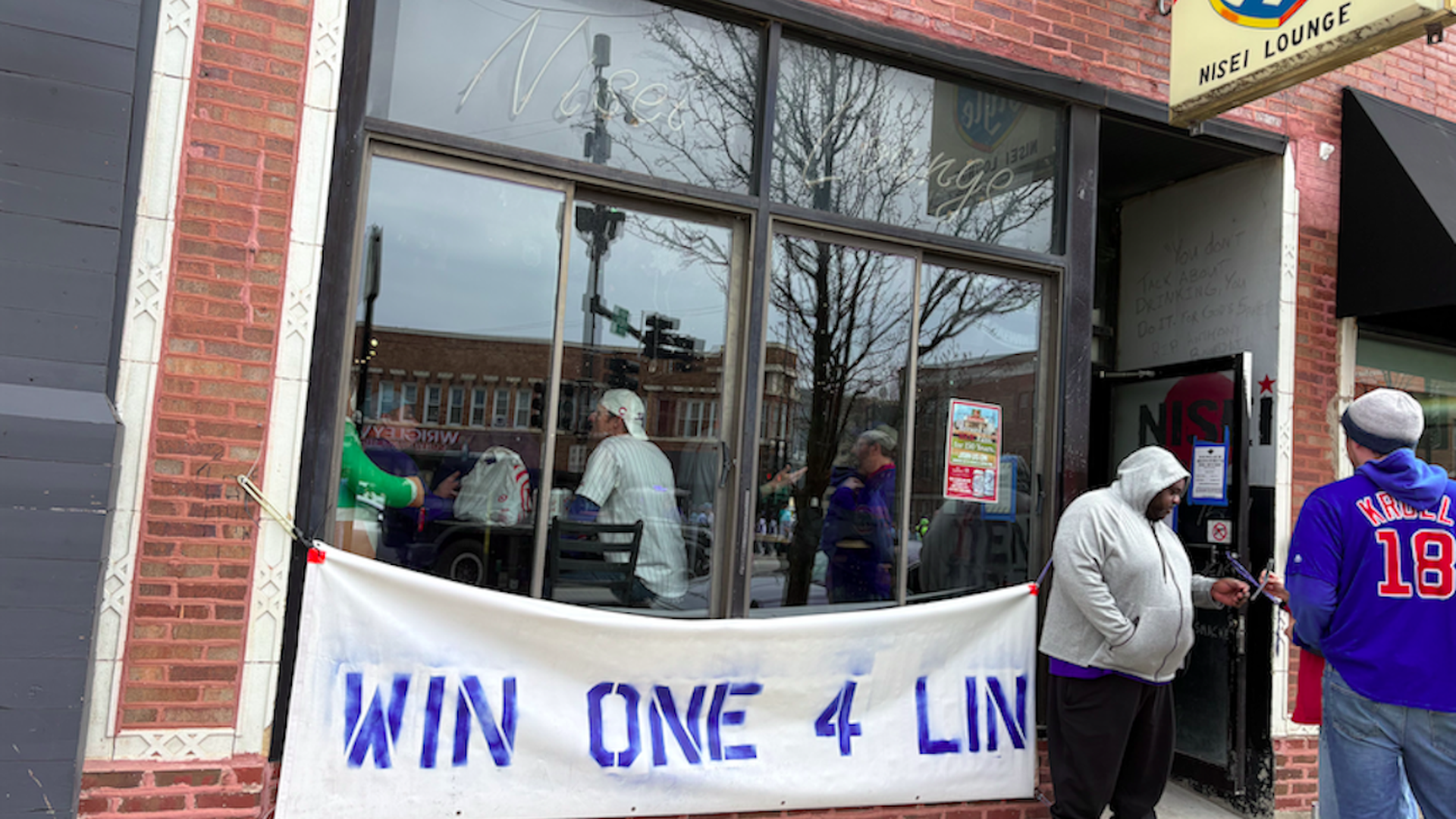 Brick storefront with large glass windows; a white banner in blue letters spans the front. Several people in hoodies stand by the entrance, including a man in a blue jersey with orange numbers.