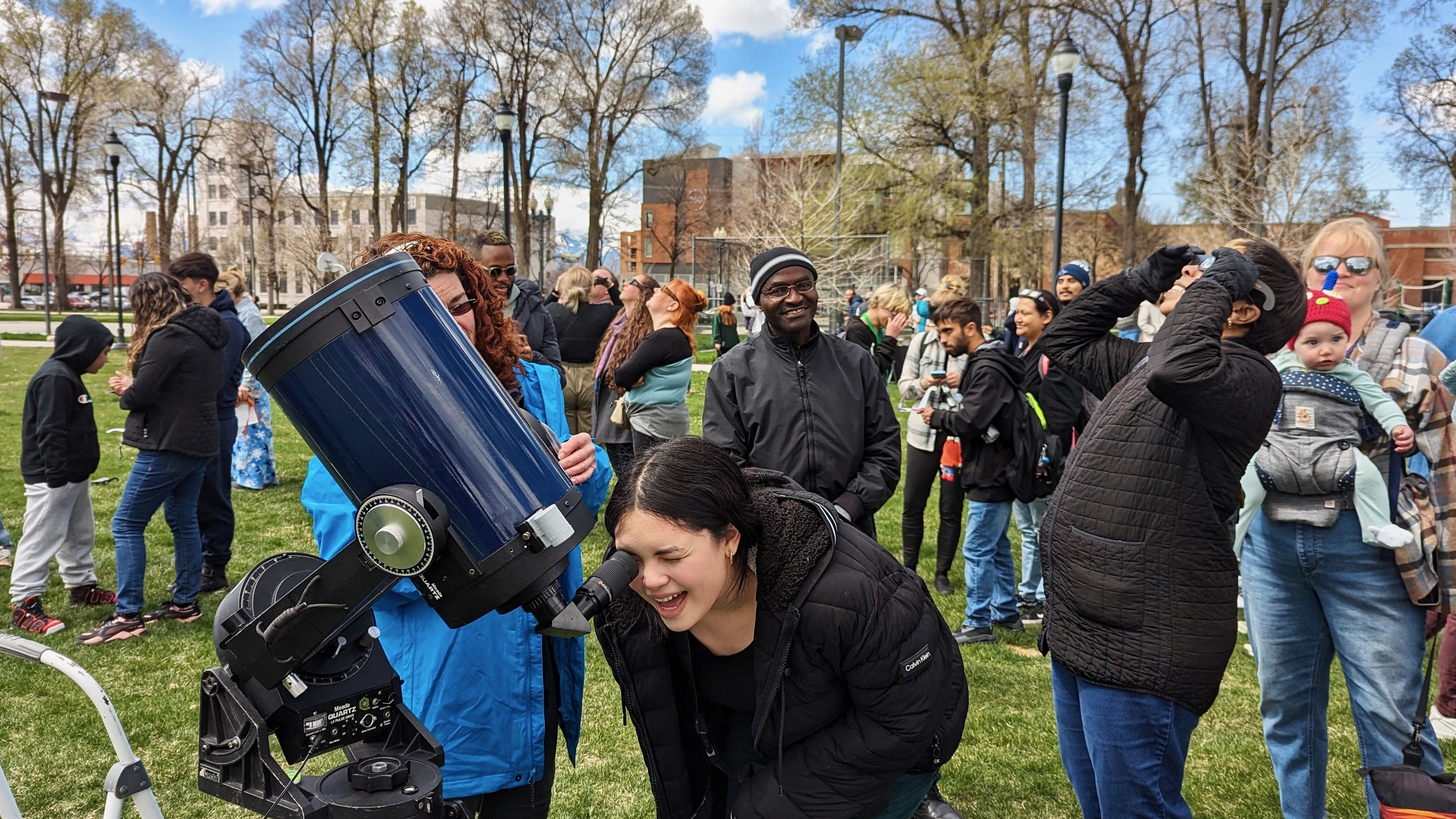 A girl smiles while looking into a large telescope in a park.