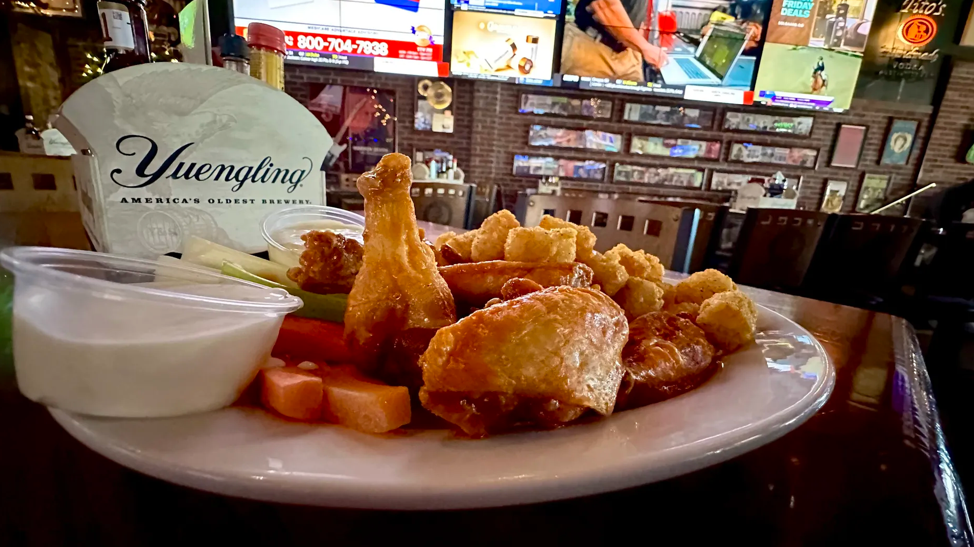 Plate of fried chicken pieces and tater tots with a white dipping sauce on a bar table; a Yuengling beer carrier and several TVs on a brick-wall backdrop are visible.