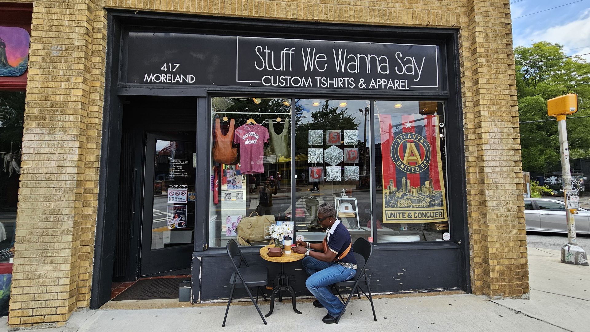 Patrice Hull sits outside her storefront at "Stuff We Wanna Say" on  Moreland Avenue. The display includes a pink T-shirt, two bags and an Atlanta United FC banner.