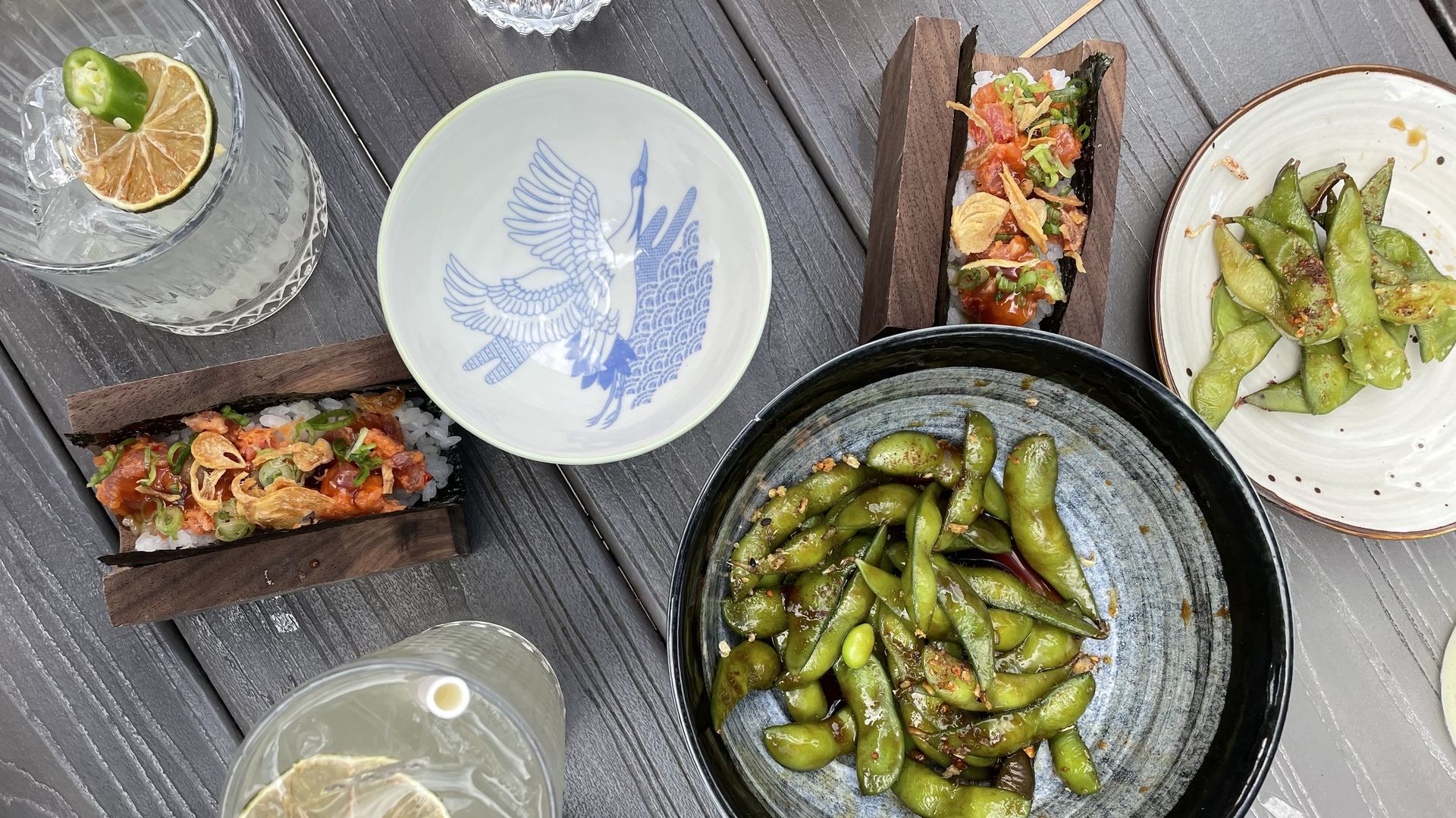 A wooden table with several dishes, including a spicy tuna roll and edamame, and two cocktails.