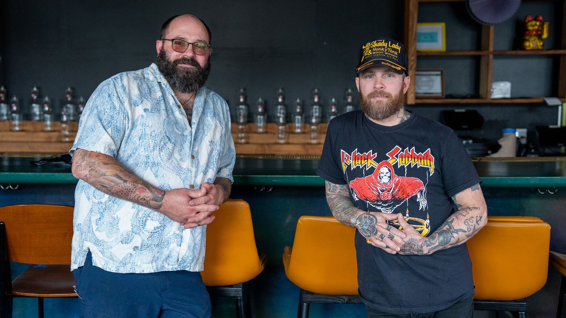 Two bearded men with tattoos stand in front of bar stools and a dark wall. One wears a blue paisley shirt and glasses, the other a Black Sabbath tee and a black cap with yellow text.