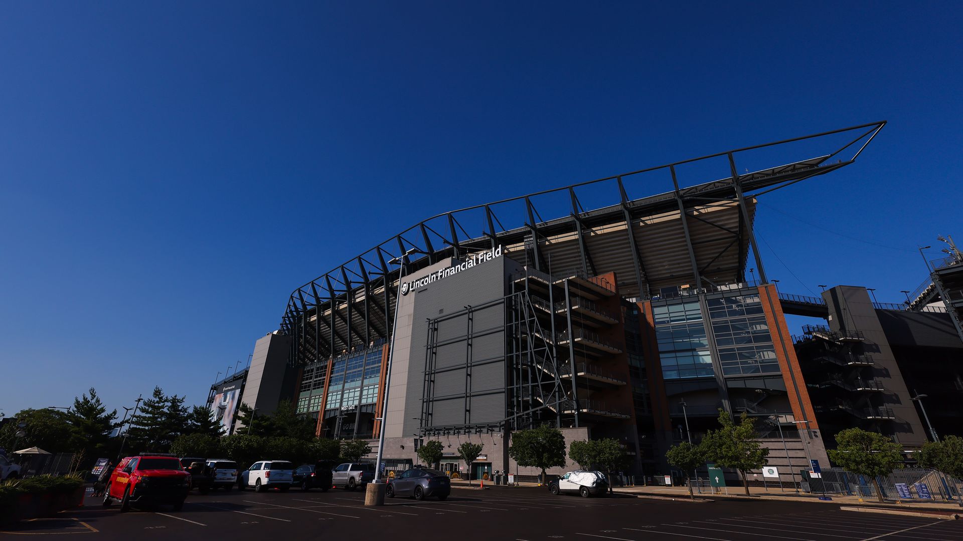 Bright blue sky above Lincoln Financial Field stadium. Exposed steel framework and brick facade, with a row of parked cars in the foreground.