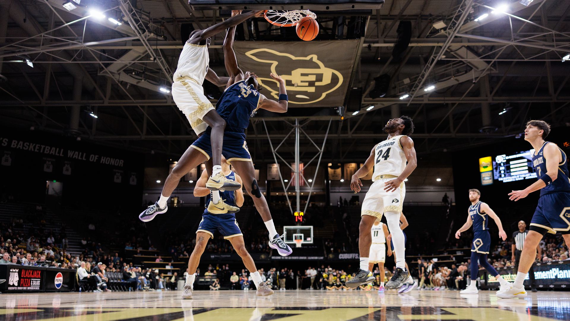 Basketball game action with player in CU jersey dunking over player in blue jersey under a hoop in an indoor stadium filled with spectators.