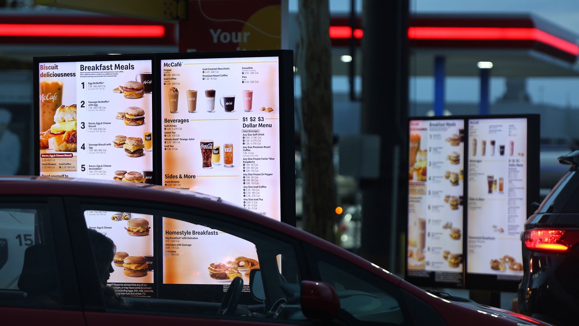 Nighttime McDonald's drive-thru with large illuminated menus displaying breakfast items and beverages; a red car is in the foreground as more menus glow in the background.