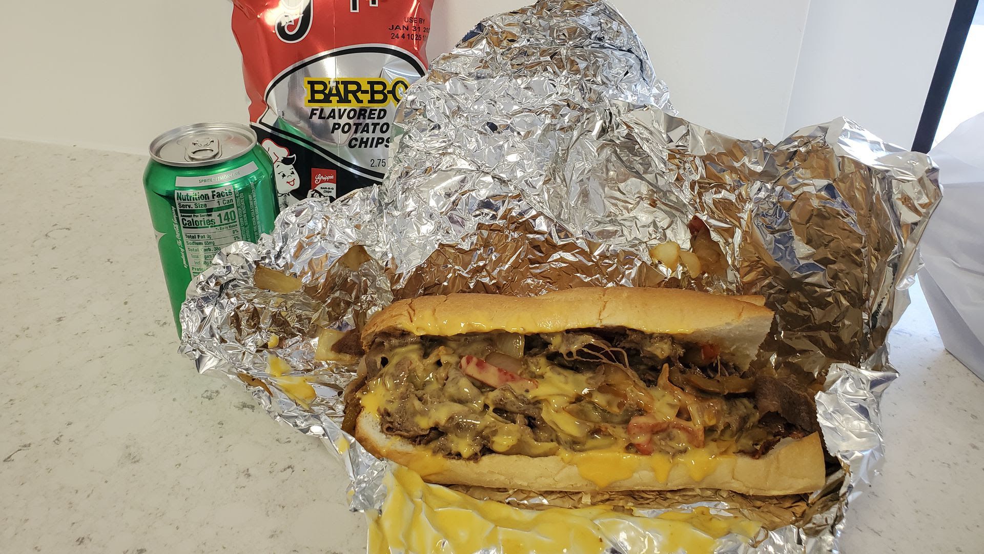 A cheesesteak wrapped in foil covered in cheese sauce, with a bag of potato chips and a can of soda next to it
