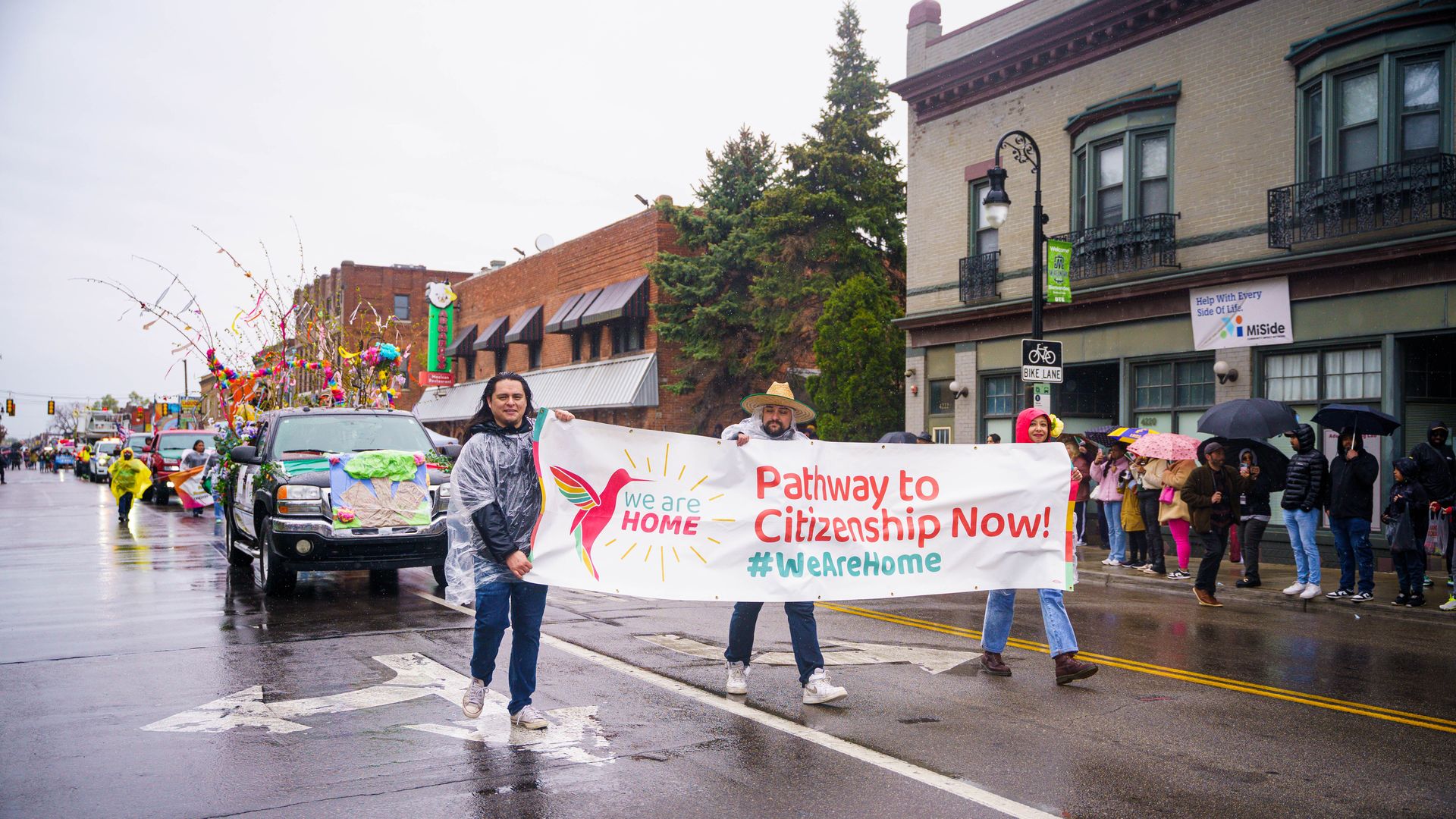 Rainy street parade with a banner that reads "Pathway to Citizenship Now!" and crowd along brick storefronts; colorful decorations on a float as marchers walk with umbrellas.