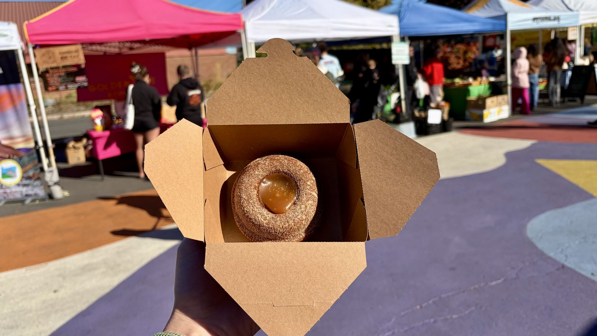 A hand holds an open cardboard box containing a glazed donut, dusted with sugar, with a caramel center. Market stalls with colorful tents and people in the background on a sunny day.