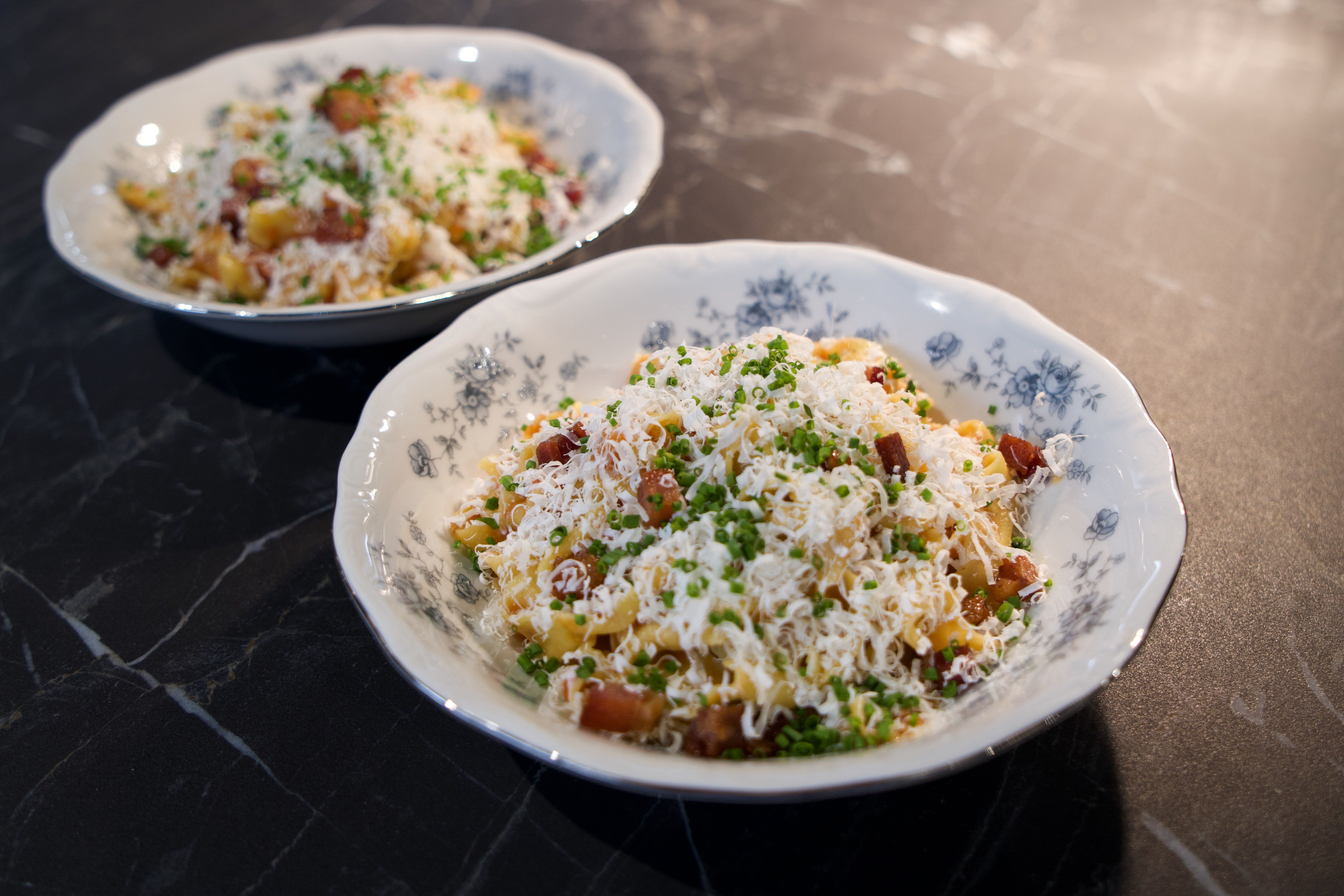 Two white bowls with blue floral designs filled with pasta topped with grated cheese, diced bacon, and chopped green herbs on a black marble surface.