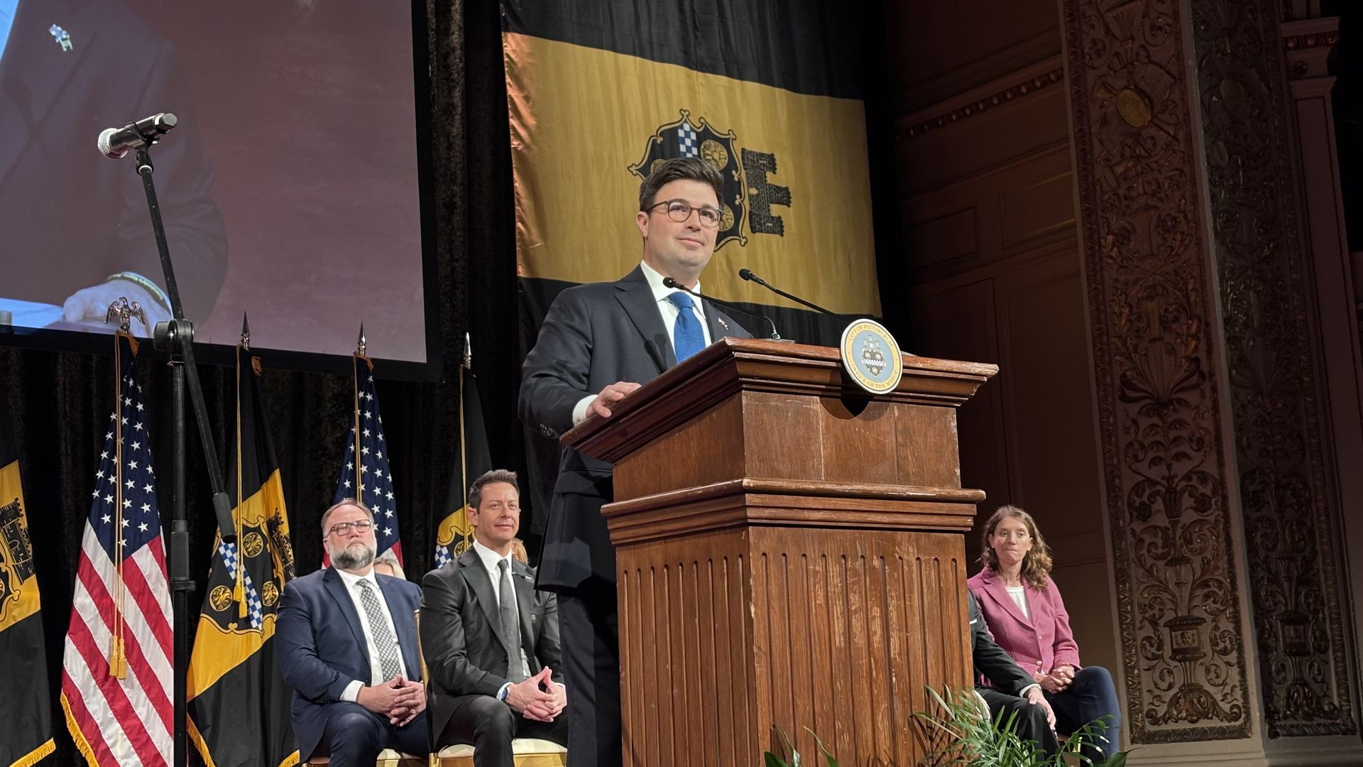 A man in a blue suit and tie speaks at a wooden podium with a City of Pittsburgh seal, with American and Pittsburgh flags behind and four seated people on stage.