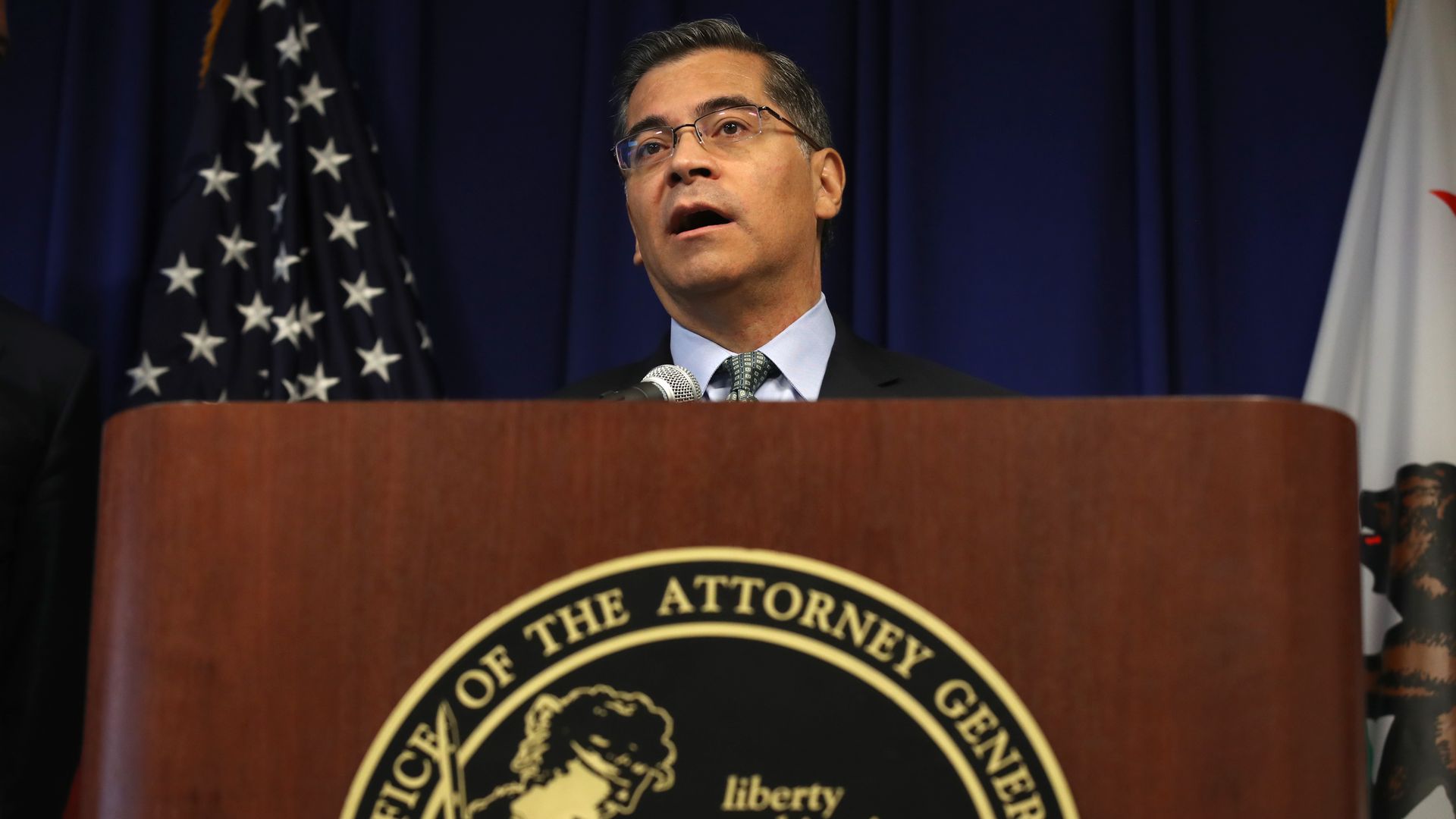 Photo of California Attorney General Xavier Becerra behind a podium next to an American flag