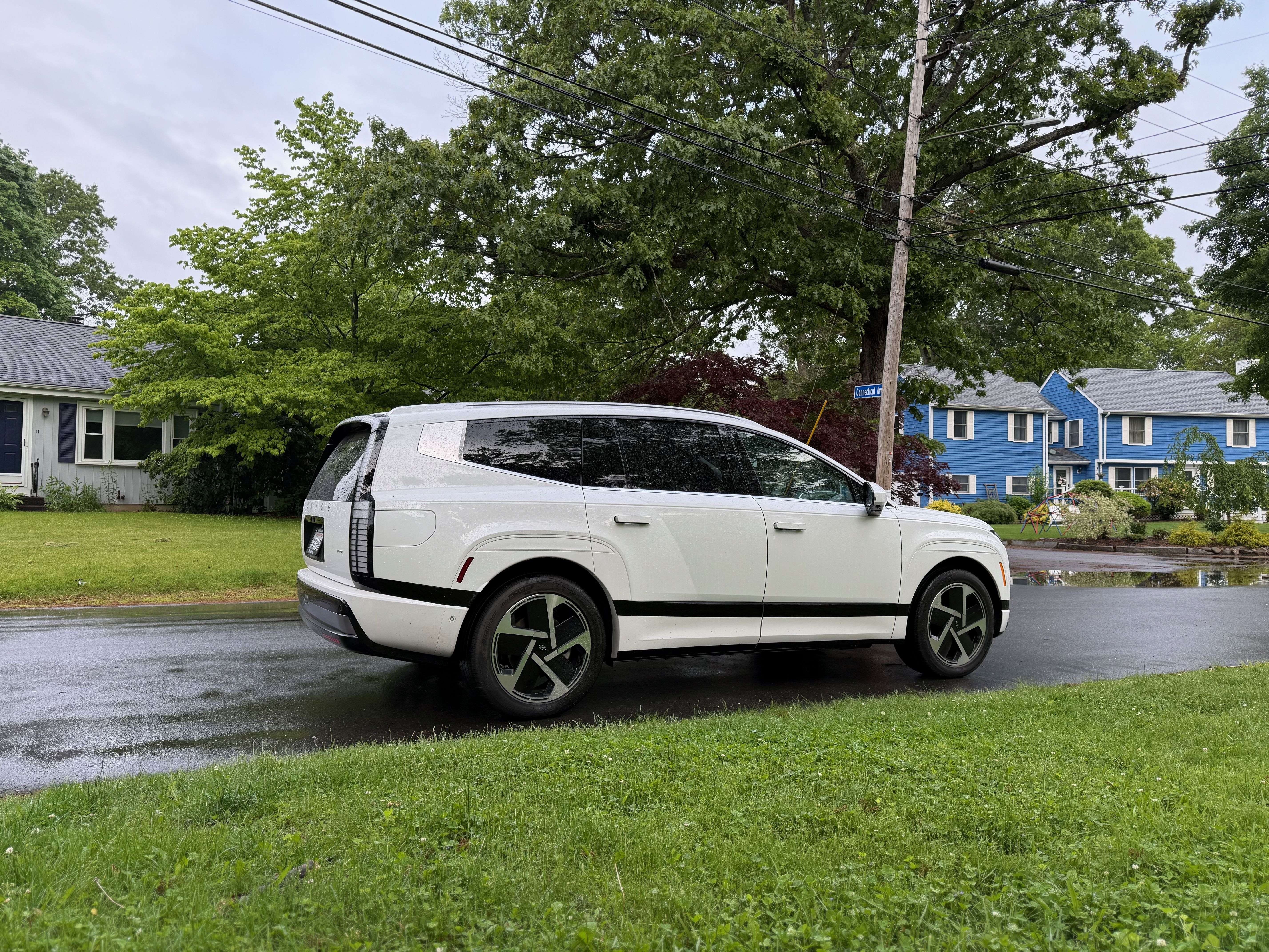 White Hyundai Ioniq 9 electric SUV parked on wet street in a residential neighborhood with green trees, grass, and blue and gray houses under cloudy sky.