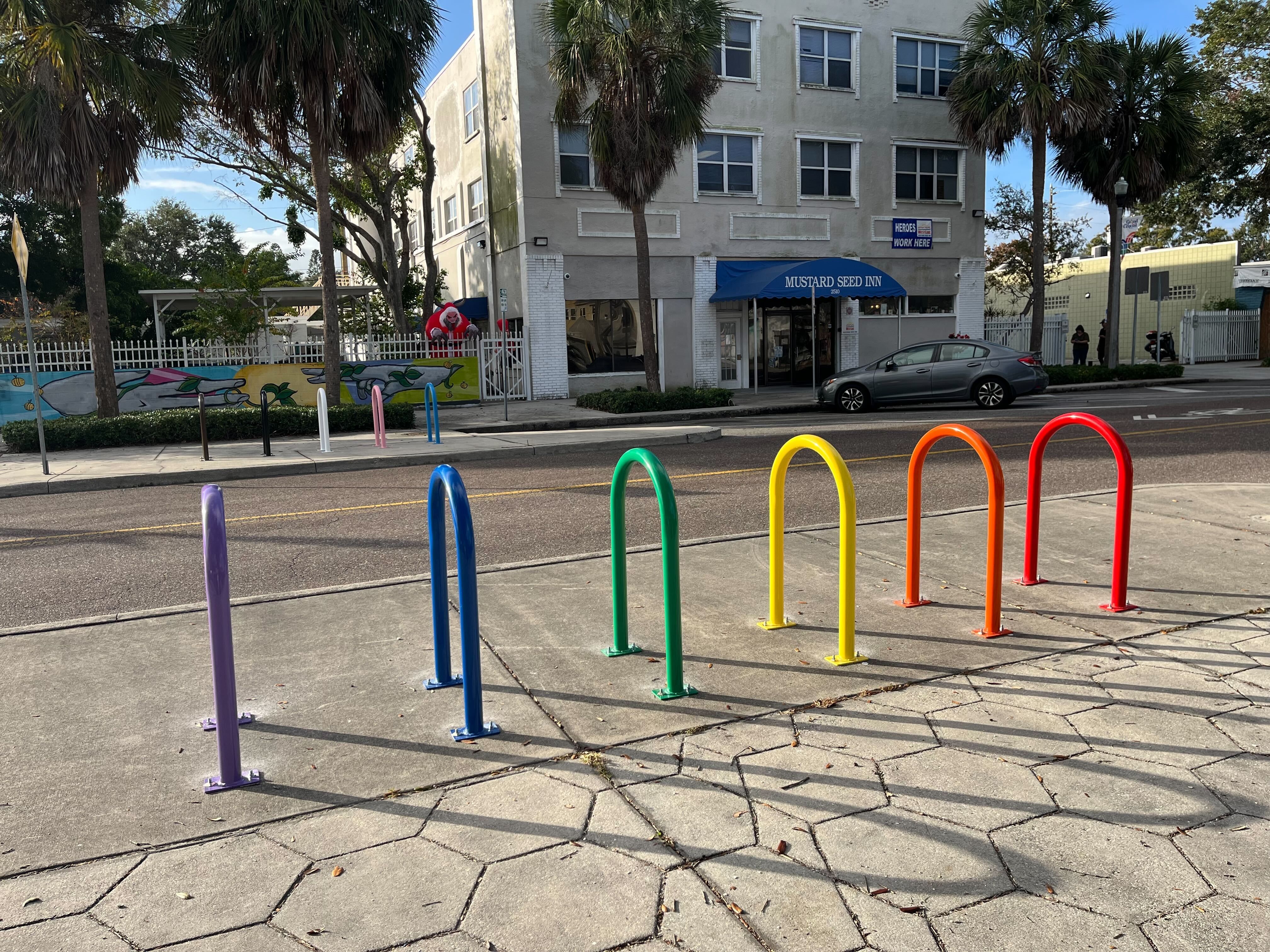 Colorful bike racks in purple, blue, green, yellow, orange, and red on concrete sidewalk across street from Mustard Seed Inn with palm trees and murals.