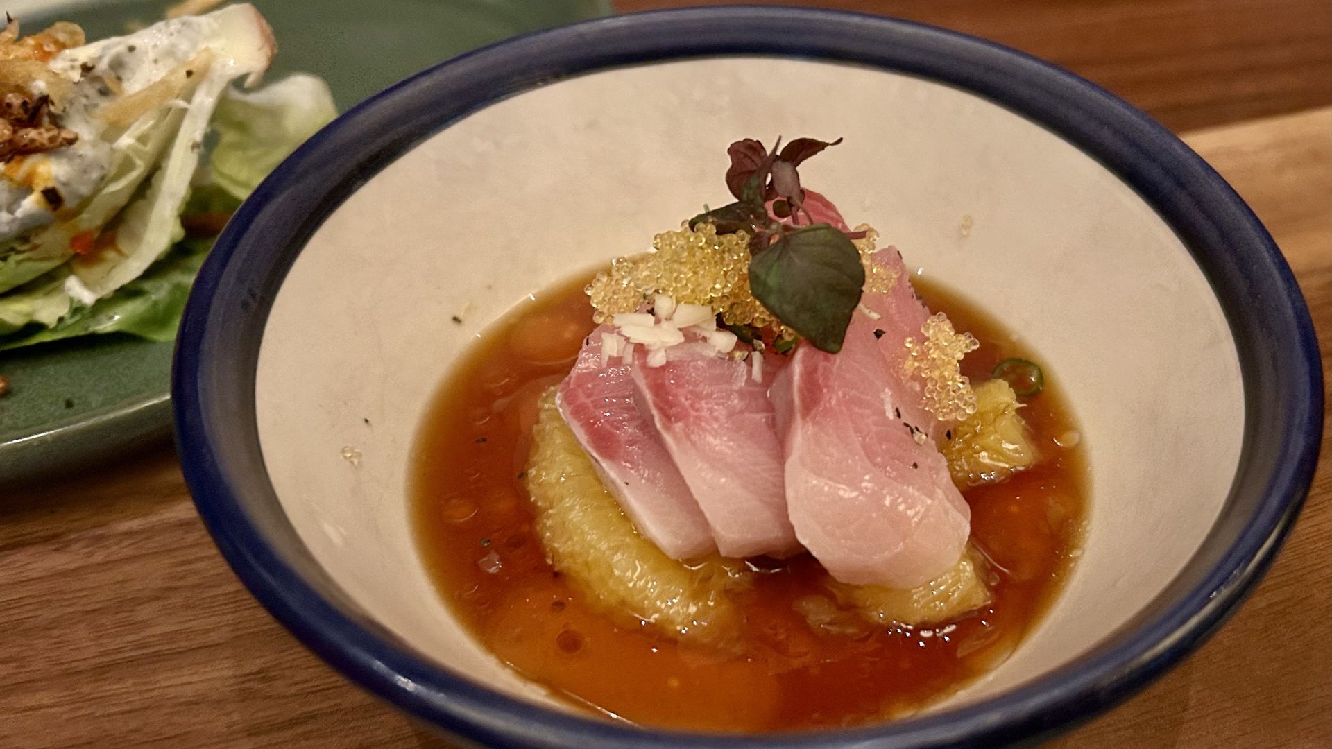 Bowl with pink sashimi on yellow fruit slices in brown sauce, garnished with small white bits, golden beads, and a small leafy herb on a wooden table.