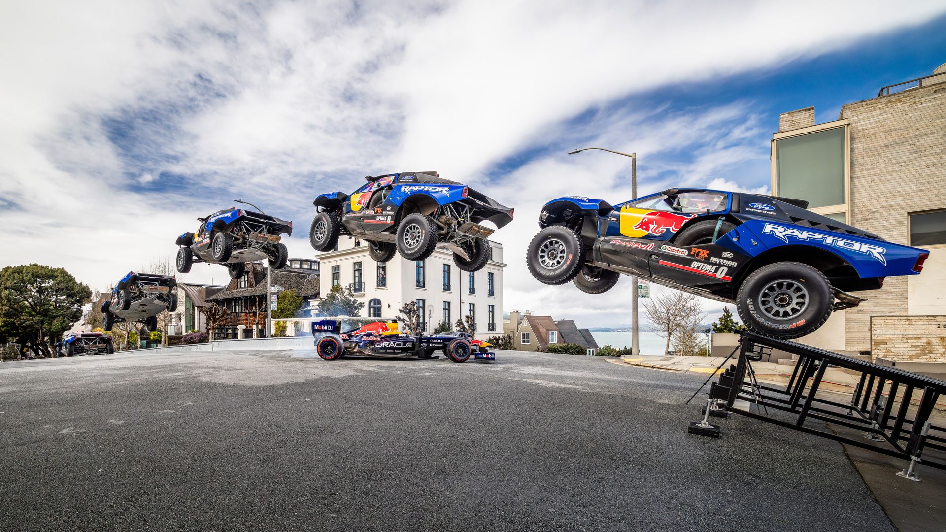 Blue and black Ford Raptor truck jumps off a ramp over a Red Bull Oracle race car on a city street with houses and trees under a cloudy sky.
