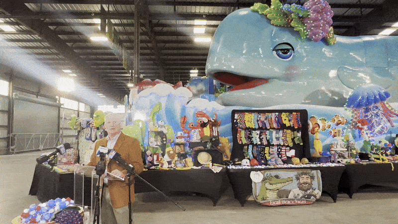 Image shows a man talking in front of a Mardi Gras float that looks like a baby whale.