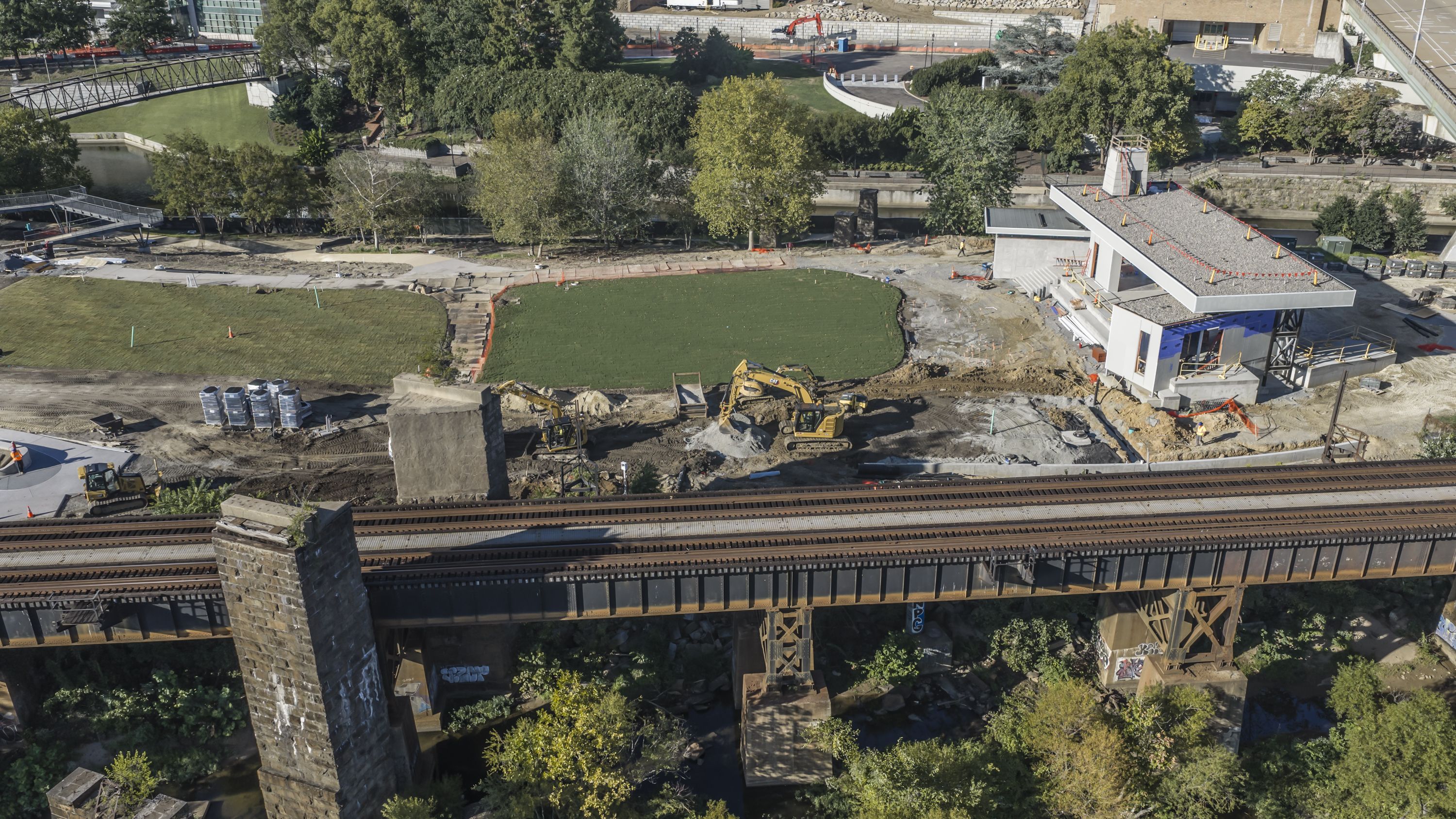 Aerial view of a riverfront construction site: excavators and dirt around a new building with a gravel roof; a grassy area, trees, orange safety cones, and a railway bridge in the foreground.
