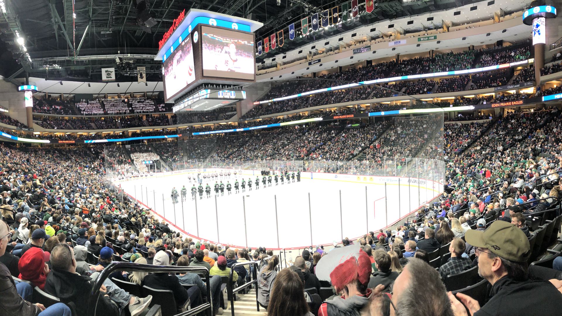 Full arena crowd watches two lined-up hockey teams on ice rink under bright lights and large overhead scoreboard inside Xcel Energy Center.