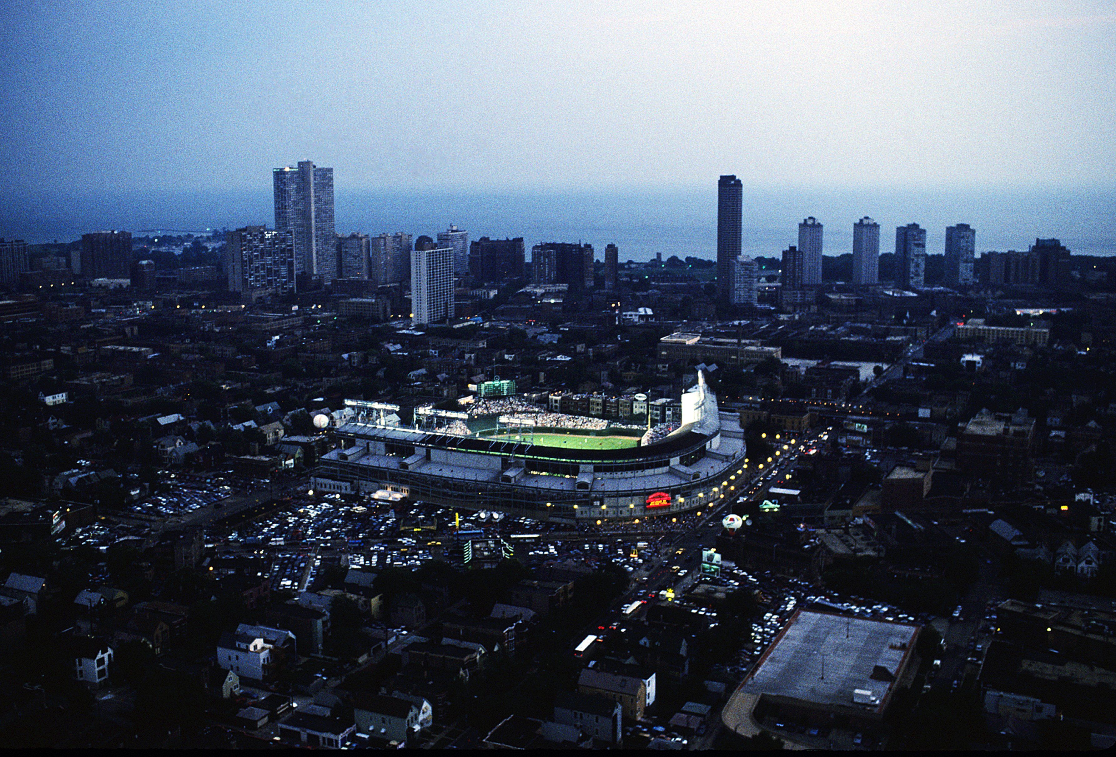 Aerial evening view of a lit sports stadium in a city with surrounding buildings and streets, under a dim blue sky near a body of water.
