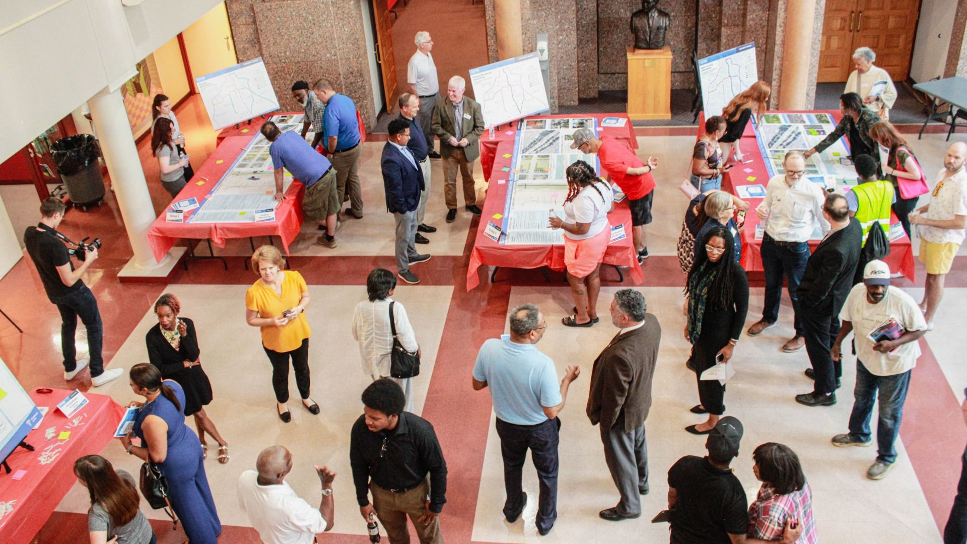 A spacious indoor lobby where a crowd browses large display boards on red tablecloths; a camera operator films left; groups converse near maps and posters, with a bust statue in the back.