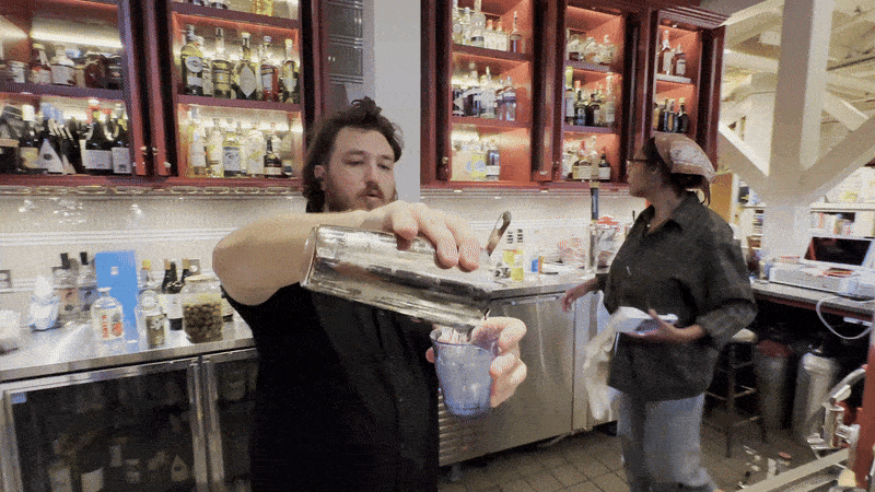Bartender with brown hair and beard pours drink from metal shaker into glass behind bar with shelves of liquor bottles. Woman in black and headscarf moves nearby in bar setting.