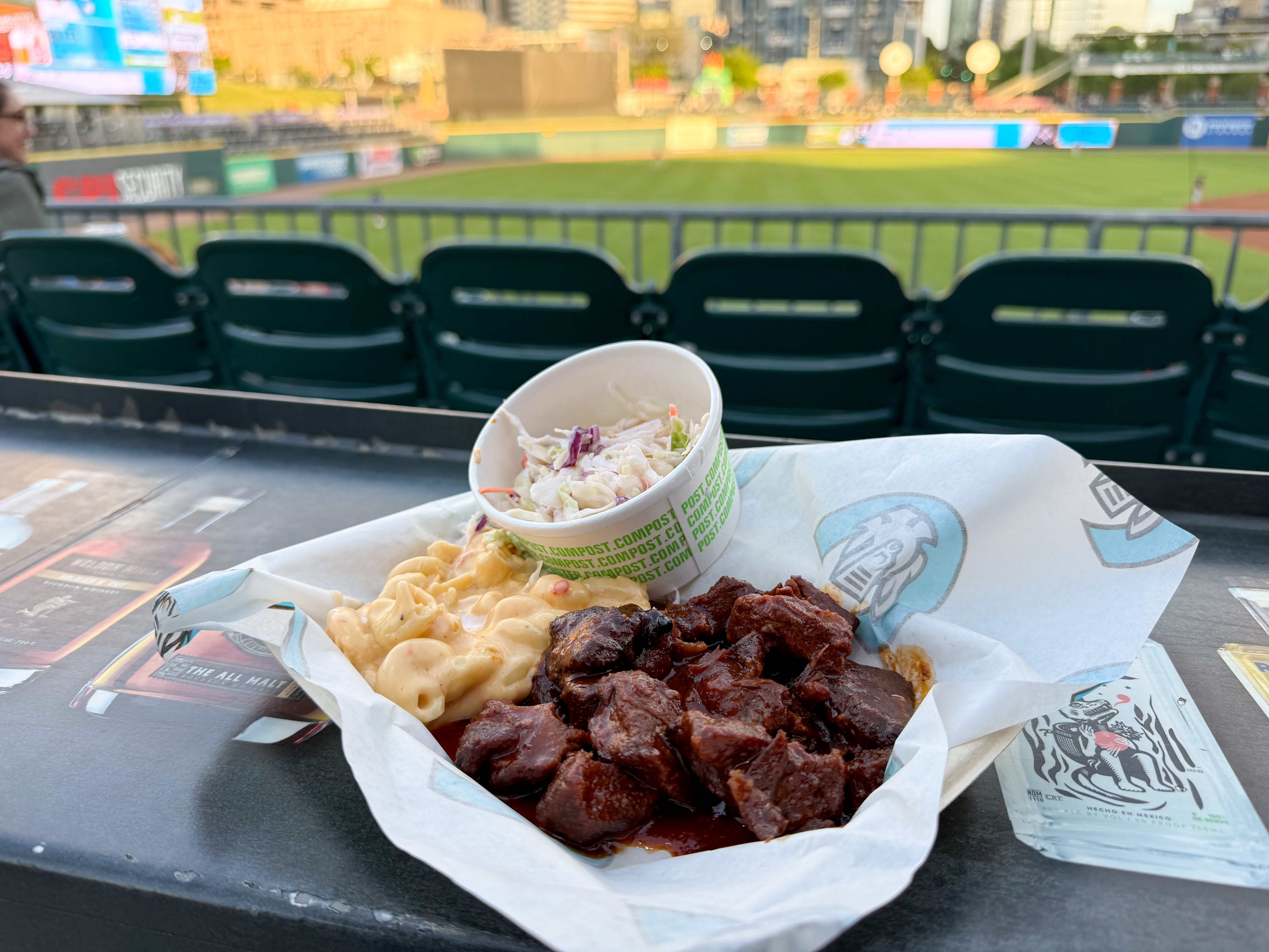 A tray of stadium food sits on a table: creamy macaroni and cheese, dark barbecued meat in sauce, and a cup of coleslaw. Empty green stadium seats and a baseball field blur in the background.