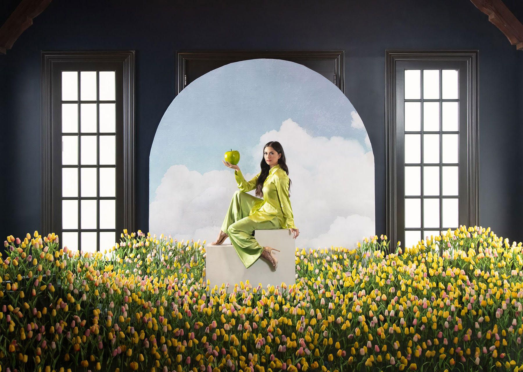 Woman in lime green satin outfit sitting on a white block holding a green apple amid a field of yellow and pink tulips, with a cloudy sky backdrop framed by two tall windows.