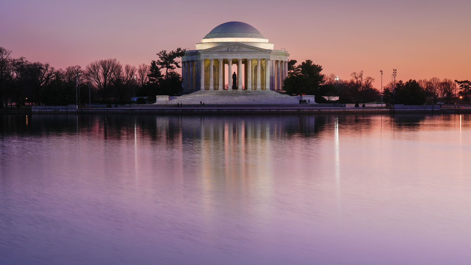 The Jefferson Memorial with its domed roof and white marble columns stands on steps by a calm reflecting pool at sunset. Silhouetted trees line the shore as pink and orange sky glows.
