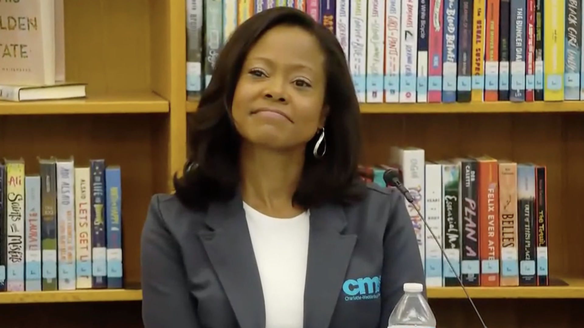 A woman with shoulder-length dark hair in a gray blazer and white blouse sits at a table in a library, smiling slightly. Colorful books line the shelves behind her, and a water bottle rests on the table.