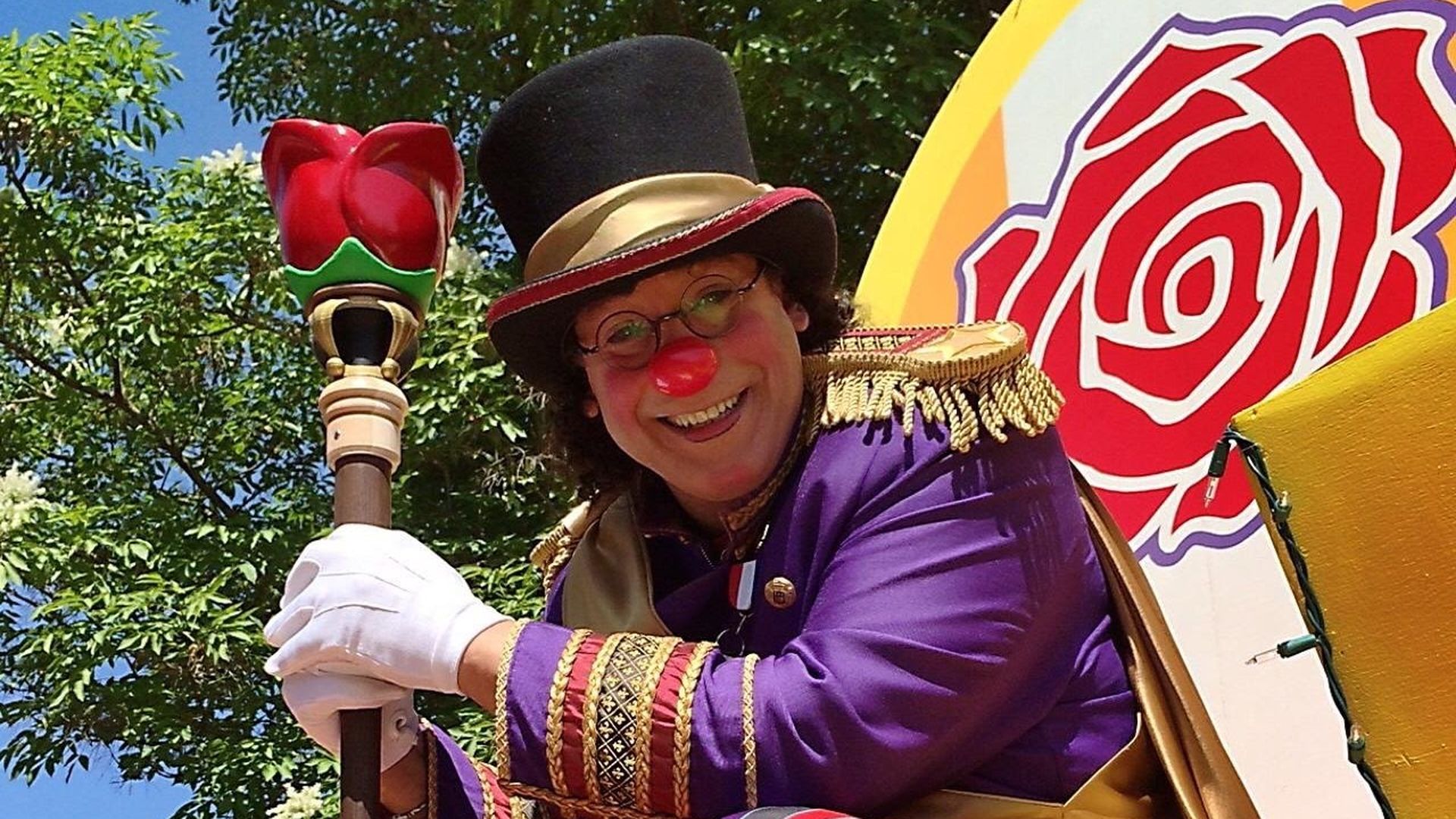 A clown wearing a red nose, black top hat, white glove and a purple jacket smiles from a float in a parade.
