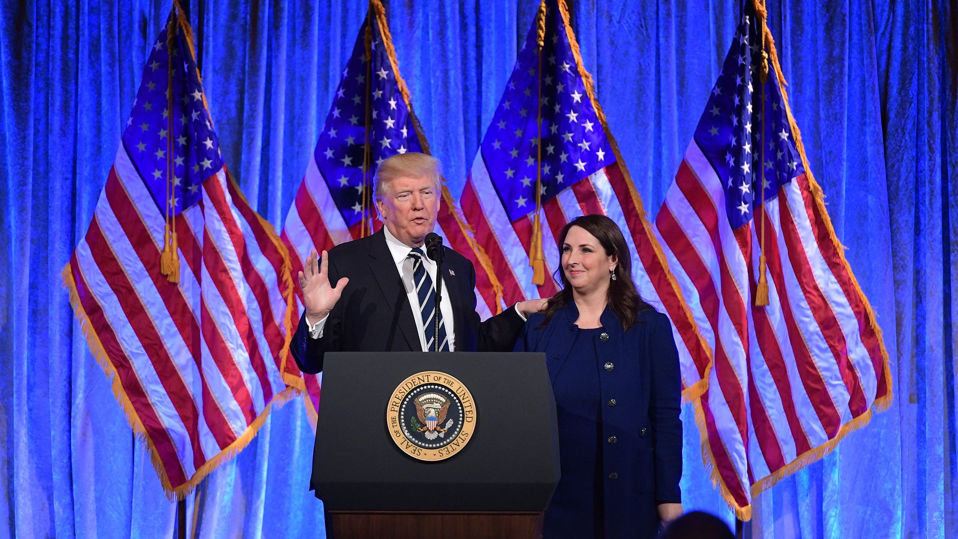 President Trump and RNC Chairwoman Ronna McDaniel in front of a blue curtain and a line of American flags. 