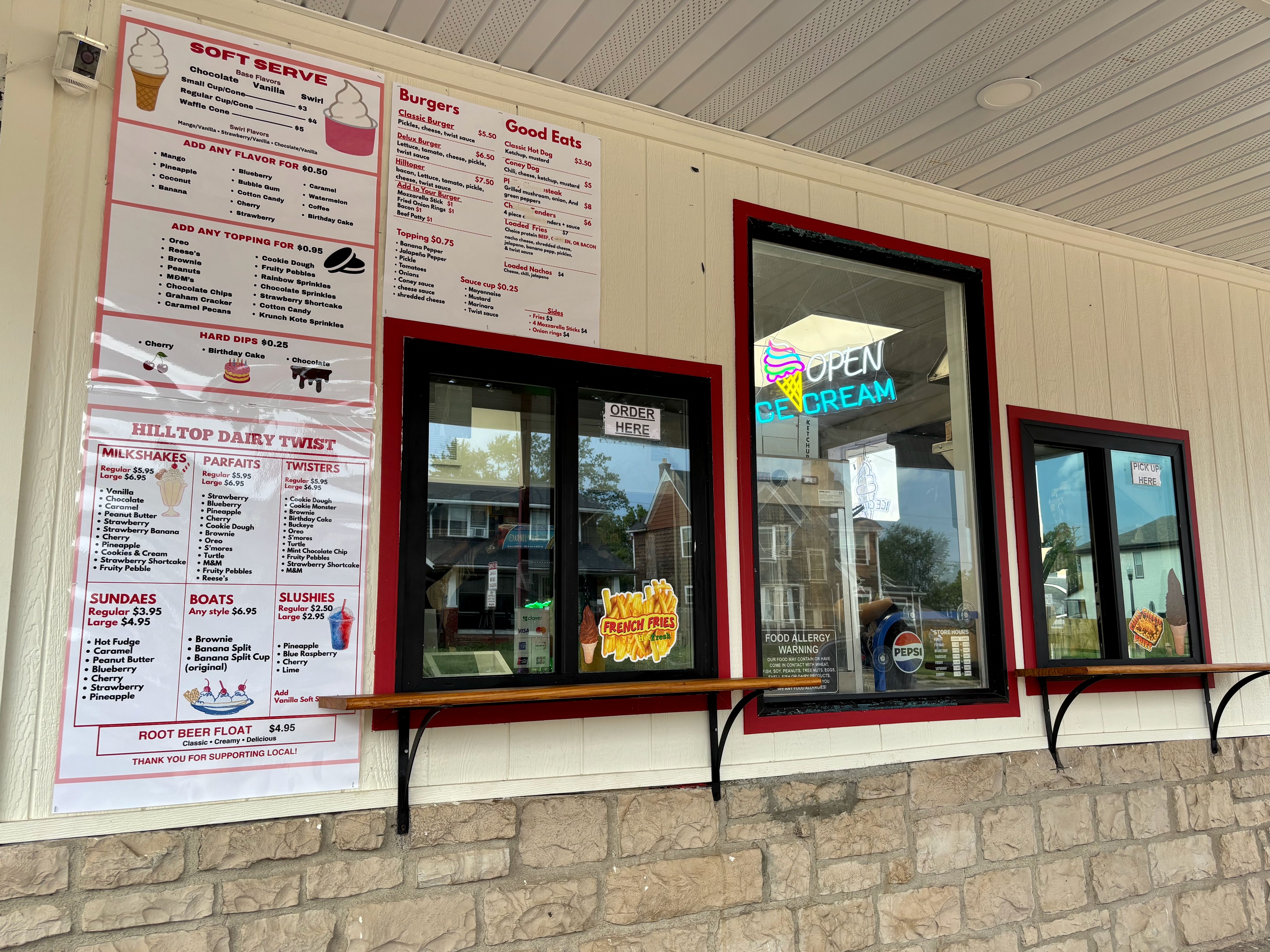 Exterior of an ice-cream shop with large wall menus for soft serve and toppings, a neon "OPEN ICE CREAM" sign in the window, a security camera on the left, beige siding, and a stone base.