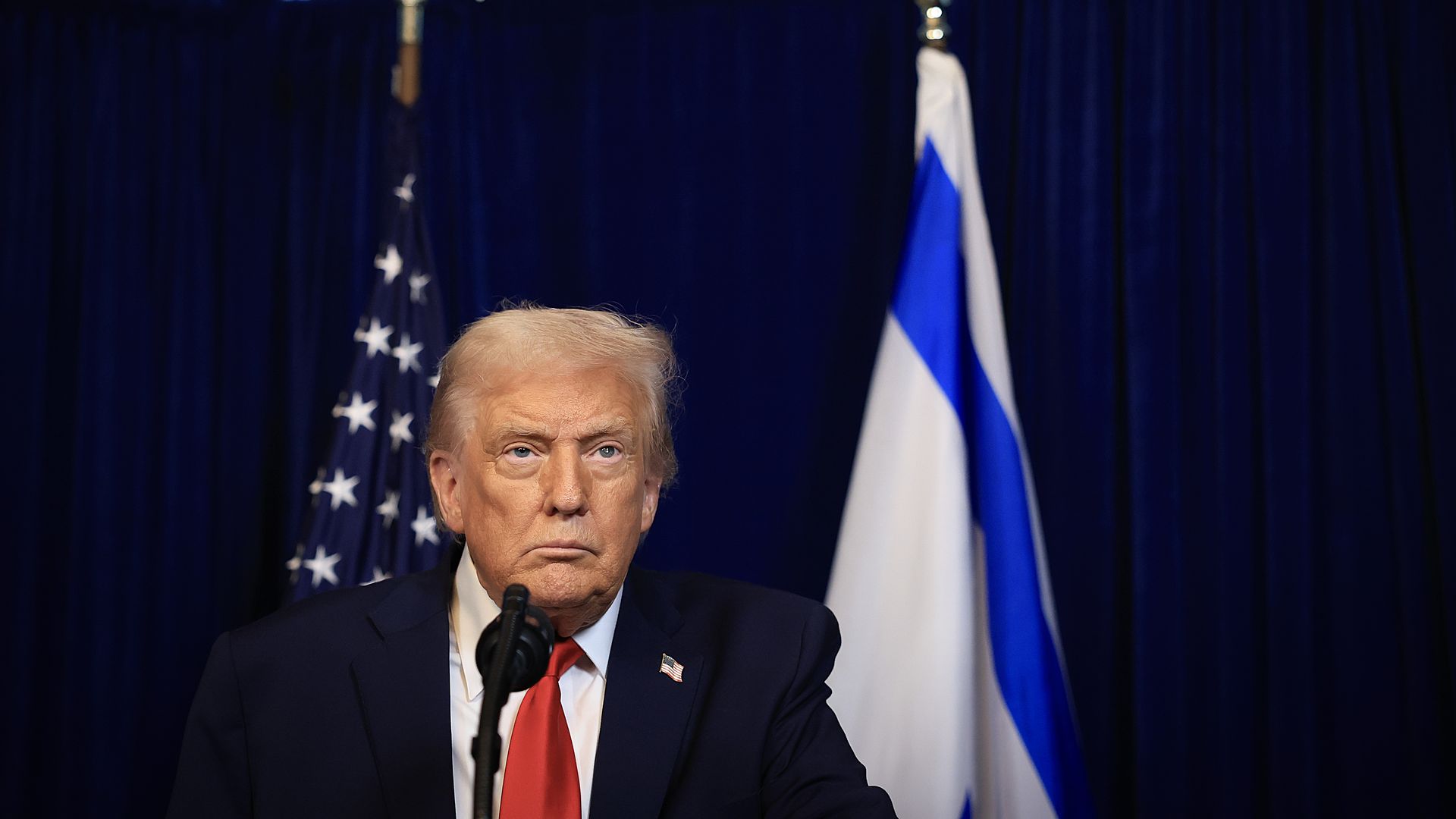 A gray-haired, fair-skinned but orange-tanned, graying President Trump, wearing a navy jacket with a US flag pin at the top of his left lapel, white shirt and red tie, stares off into the distance while sitting at a black mic in front of a US flag (L) and an Israeli flag, in front of blue curtains.