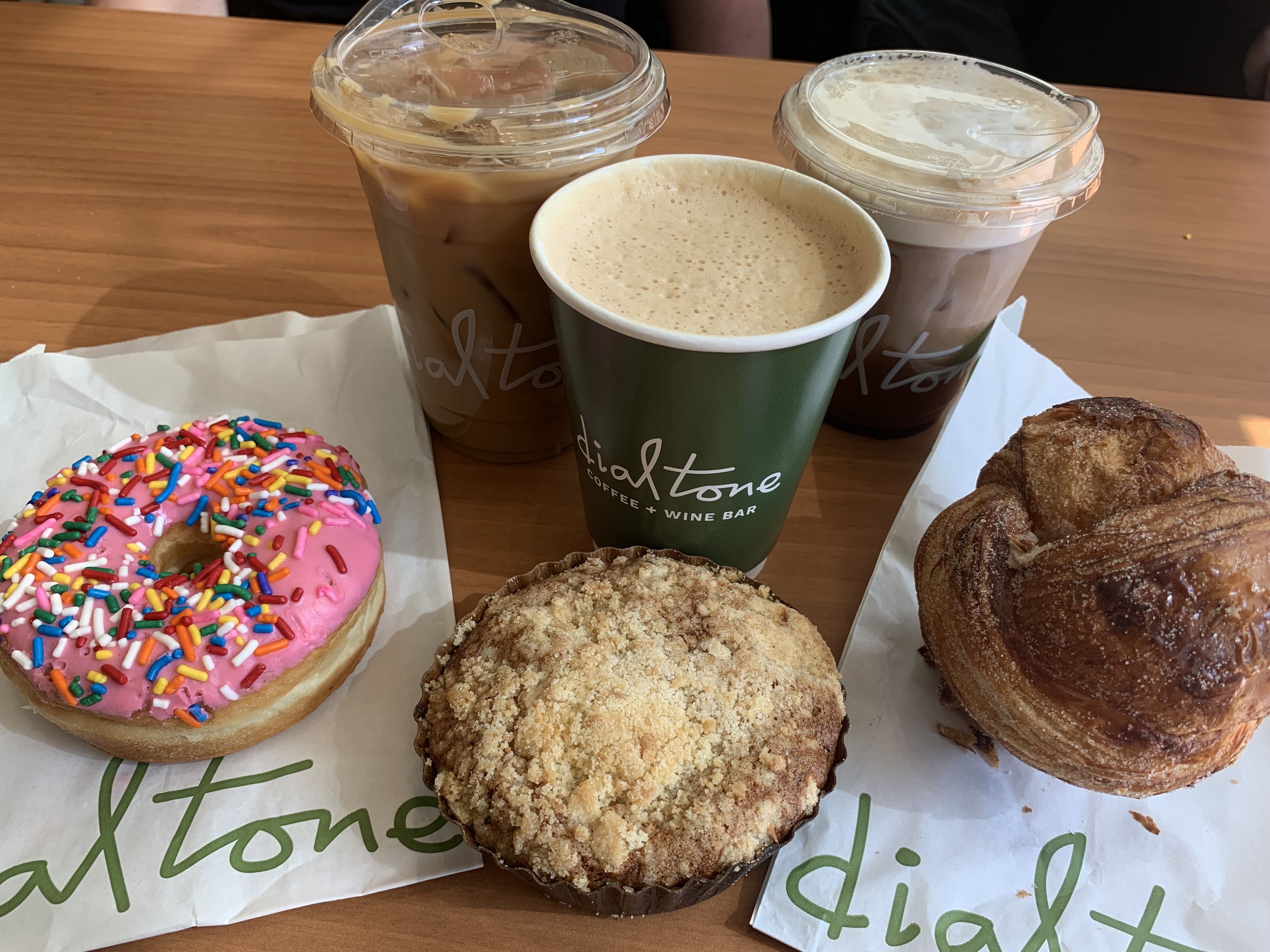 Three coffee drinks including iced and hot in cups labeled "dial tone" on a wooden table, accompanied by a pink-frosted donut with sprinkles, a crumb-topped muffin, and a croissant on paper bags.