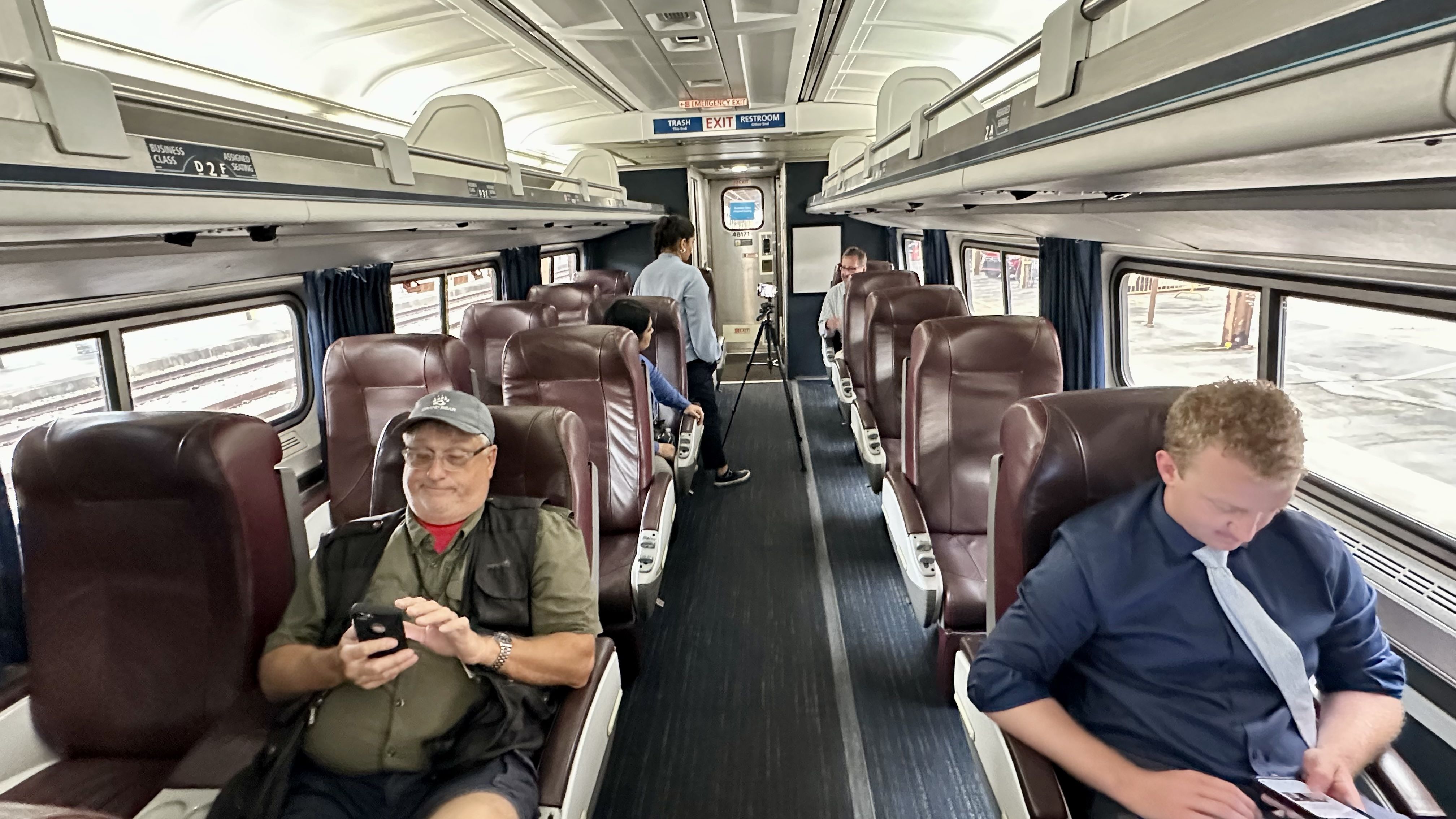 Inside a train's business class carriage with brown leather seats; five passengers engaged with phones or talking; overhead signs for trash, exit, and restroom.