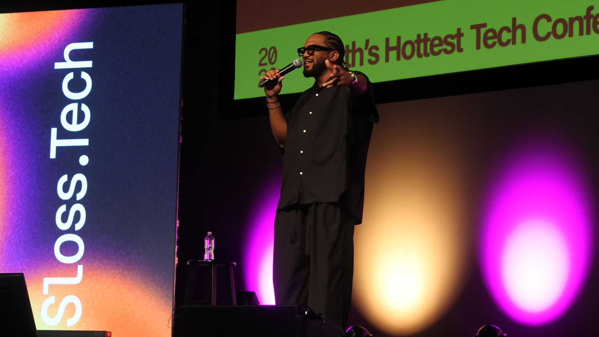 A male speaker in black clothing and glasses on a dark stage, holding a microphone and gesturing. Bright lime-green screens behind him show banners reading "Hottest Tech Conference".
