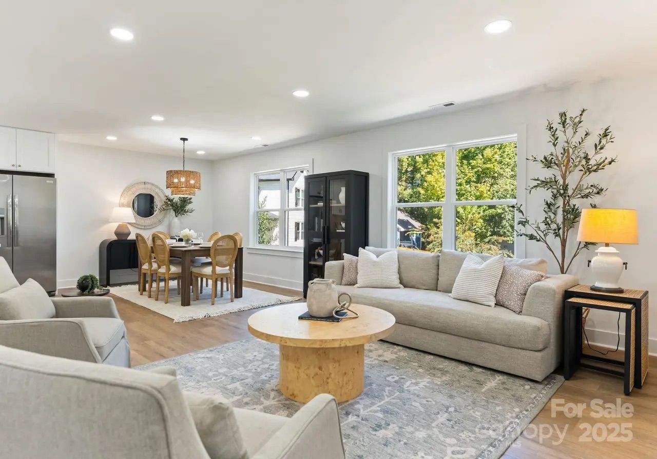 Bright living and dining area with beige sofa, wooden round coffee table, two armchairs, dining table with six chairs, large windows showing green trees, and modern decor with plants and lamps.