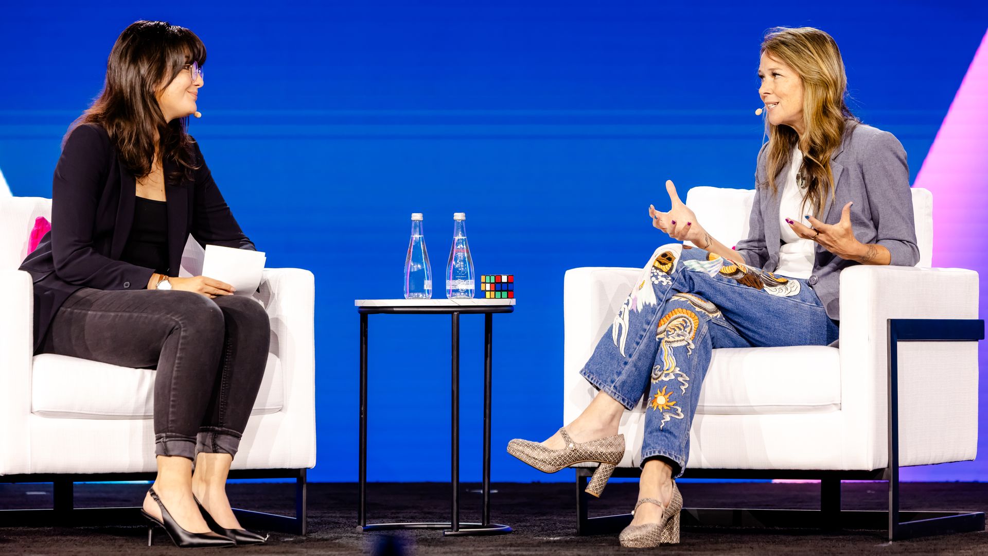 Two women seated in white chairs on a stage with blue background, engaged in conversation. One wears black blazer and jeans, the other colorful embroidered jeans and blazer. Table with water and Rubik's cube between them.