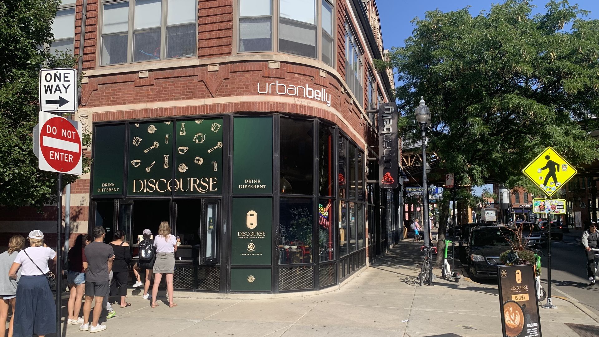 Line of people waiting outside Discourse coffee kiosk in brick urbanbelly building under clear blue sky, with street signs and yellow pedestrian crossing sign nearby.