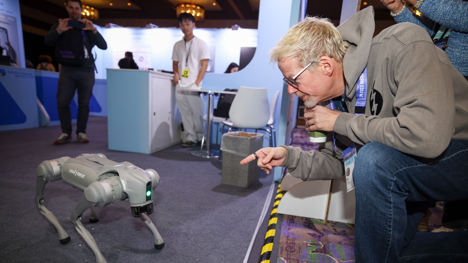 A gray four-legged robot dog with a green light on its head stands on a carpeted expo floor as a man in a gray hoodie and glasses crouches nearby, pointing and holding a cup at a trade show booth.